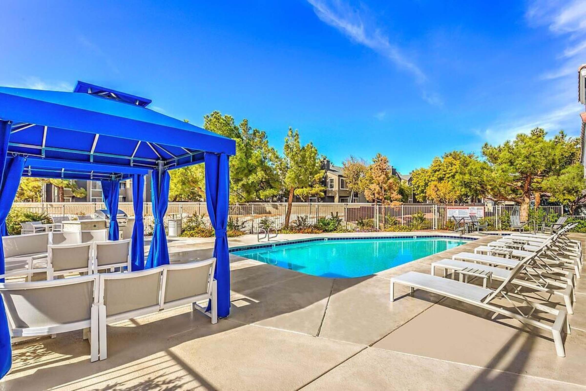 A spacious outdoor pool area featuring a clear blue pool, lounge chairs, and a shaded gazebo. Surrounding greenery and well-maintained landscaping create a relaxing atmosphere under a bright blue sky. Perfect for leisure and relaxation.
