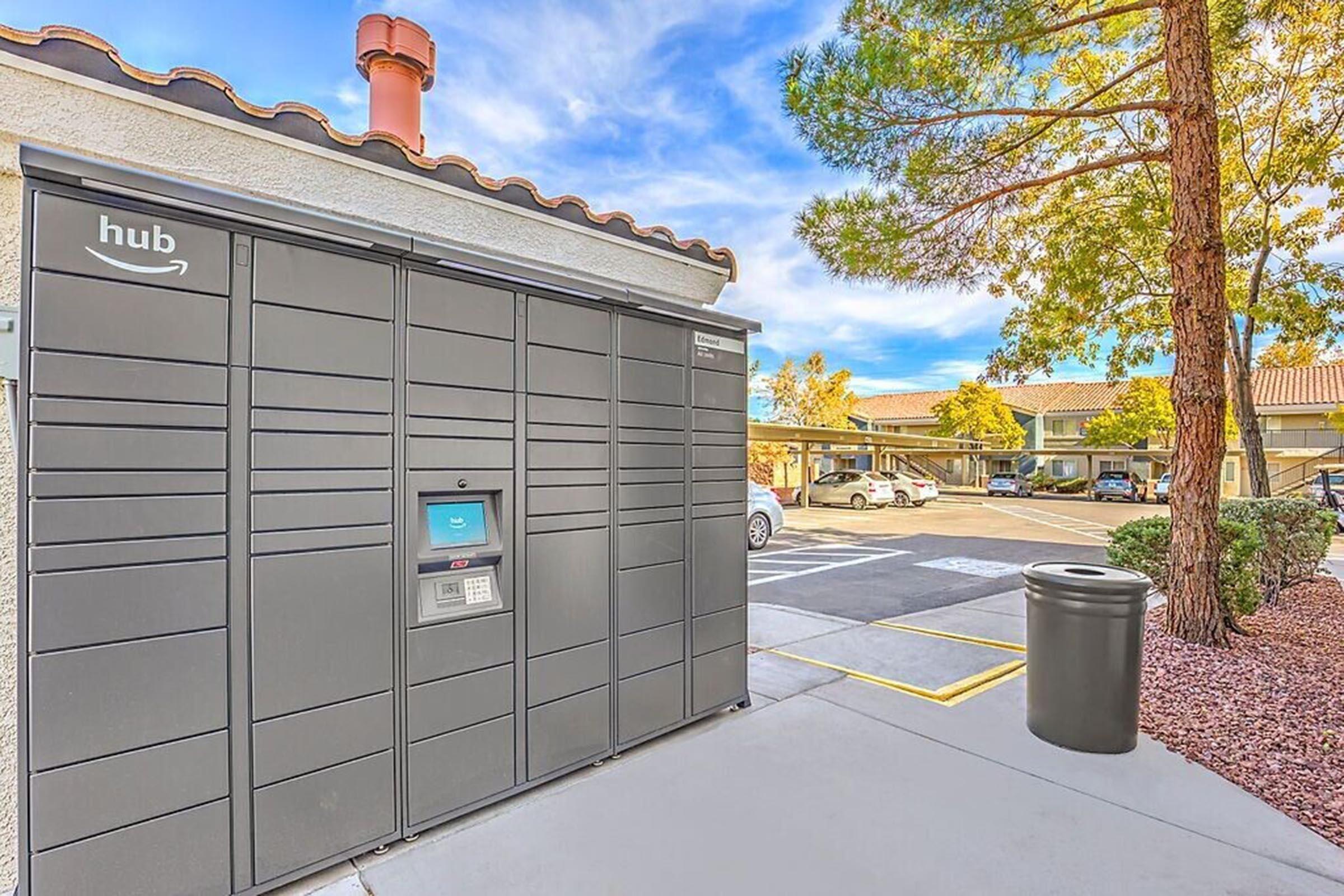 A row of Amazon Hub lockers set against a clear blue sky, surrounded by trees and a parking area. The lockers are positioned next to a trash can, with a pathway leading up to them. Nearby, cars are parked in the lot, indicating a residential or commercial complex.