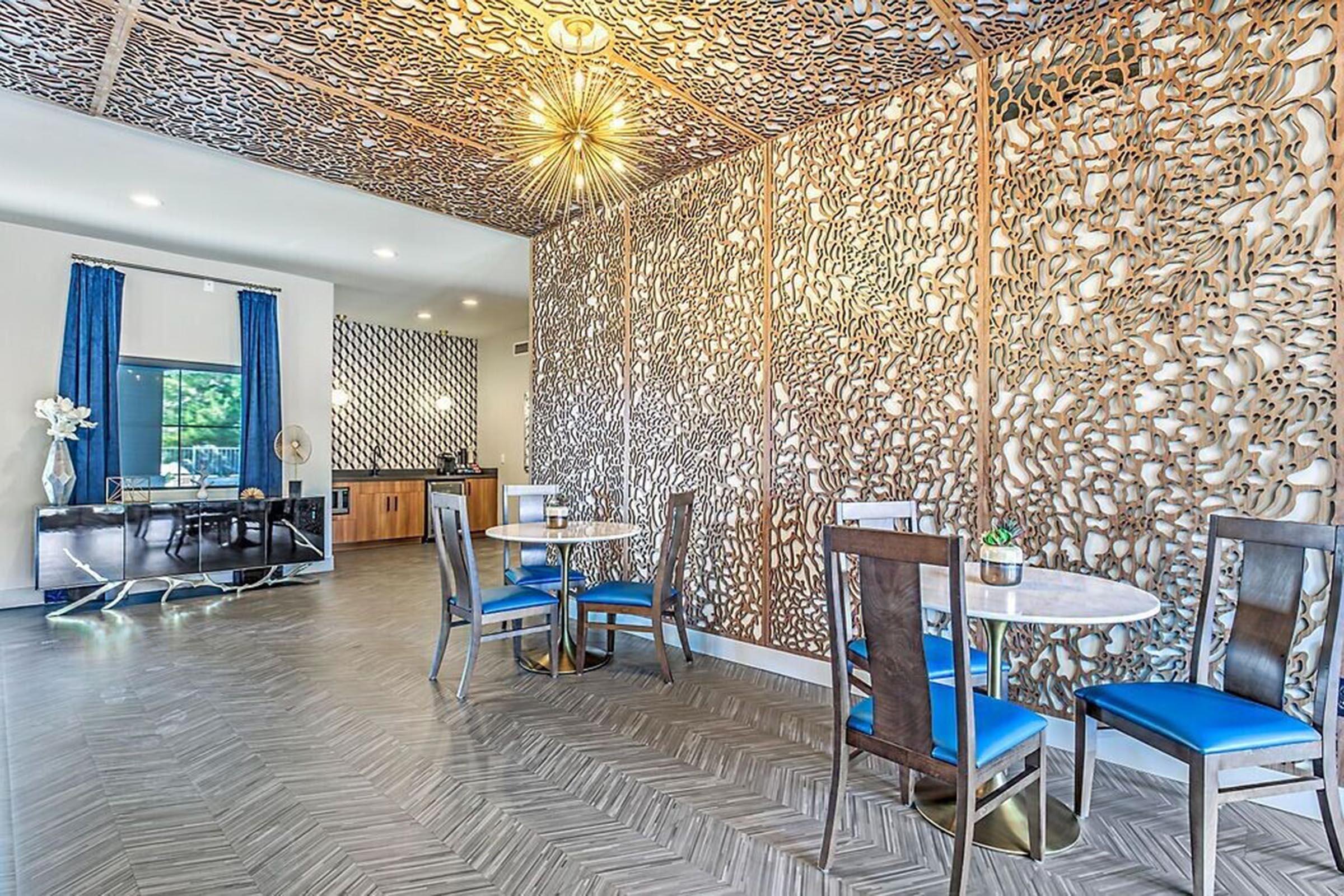 A modern dining area featuring stylish marble-topped tables with blue upholstered chairs. The wall has an intricate geometric design, and there’s a contemporary light fixture above. A kitchen area is visible in the background, with wooden cabinetry and decorative elements. Natural light brightens the space.