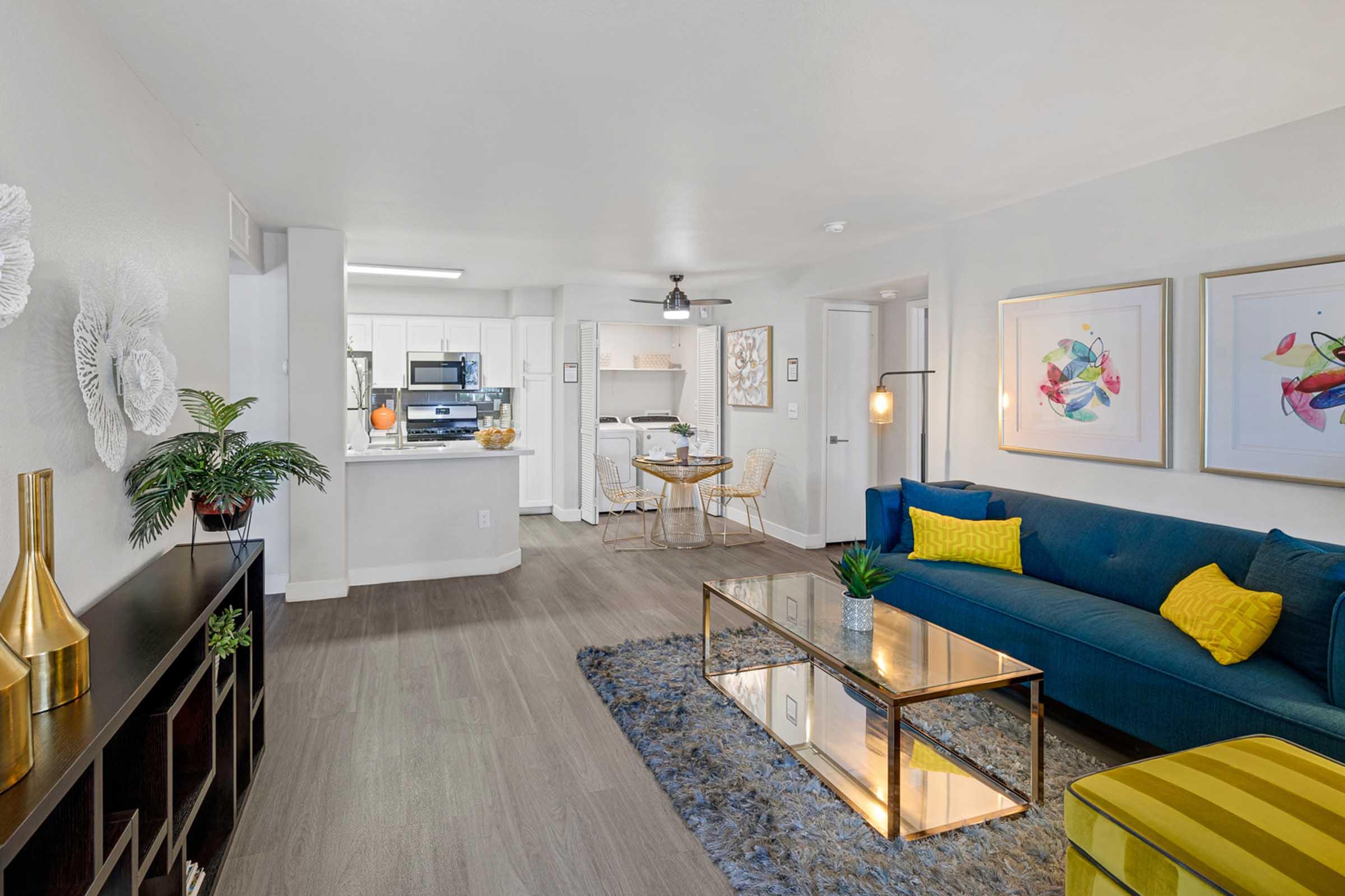 A modern living room featuring a blue sofa with decorative cushions, a glass coffee table, a stylish rug, and a plant. In the background, an open kitchen with white cabinets and appliances. Abstract art pieces are framed on the walls, and there is a dining area with a round table and chairs.