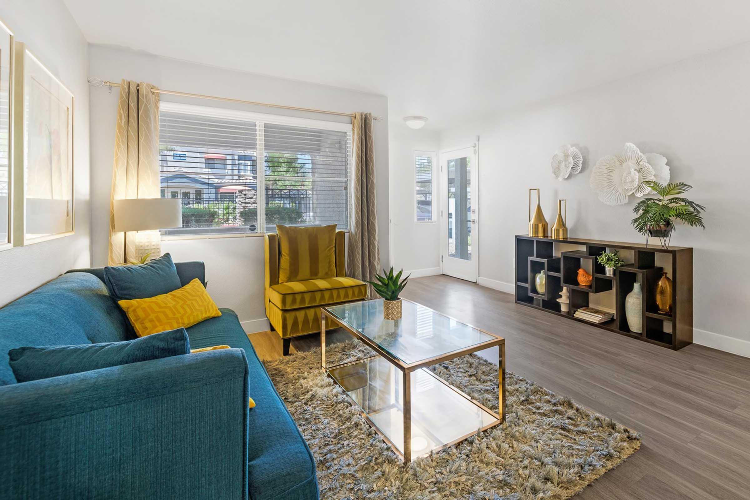 Interior of a modern living room featuring a blue couch with yellow pillows, a glass coffee table on a plush area rug, and a stylish side table with decorative items. Large window with curtains lets in natural light, enhancing the warm, inviting atmosphere.