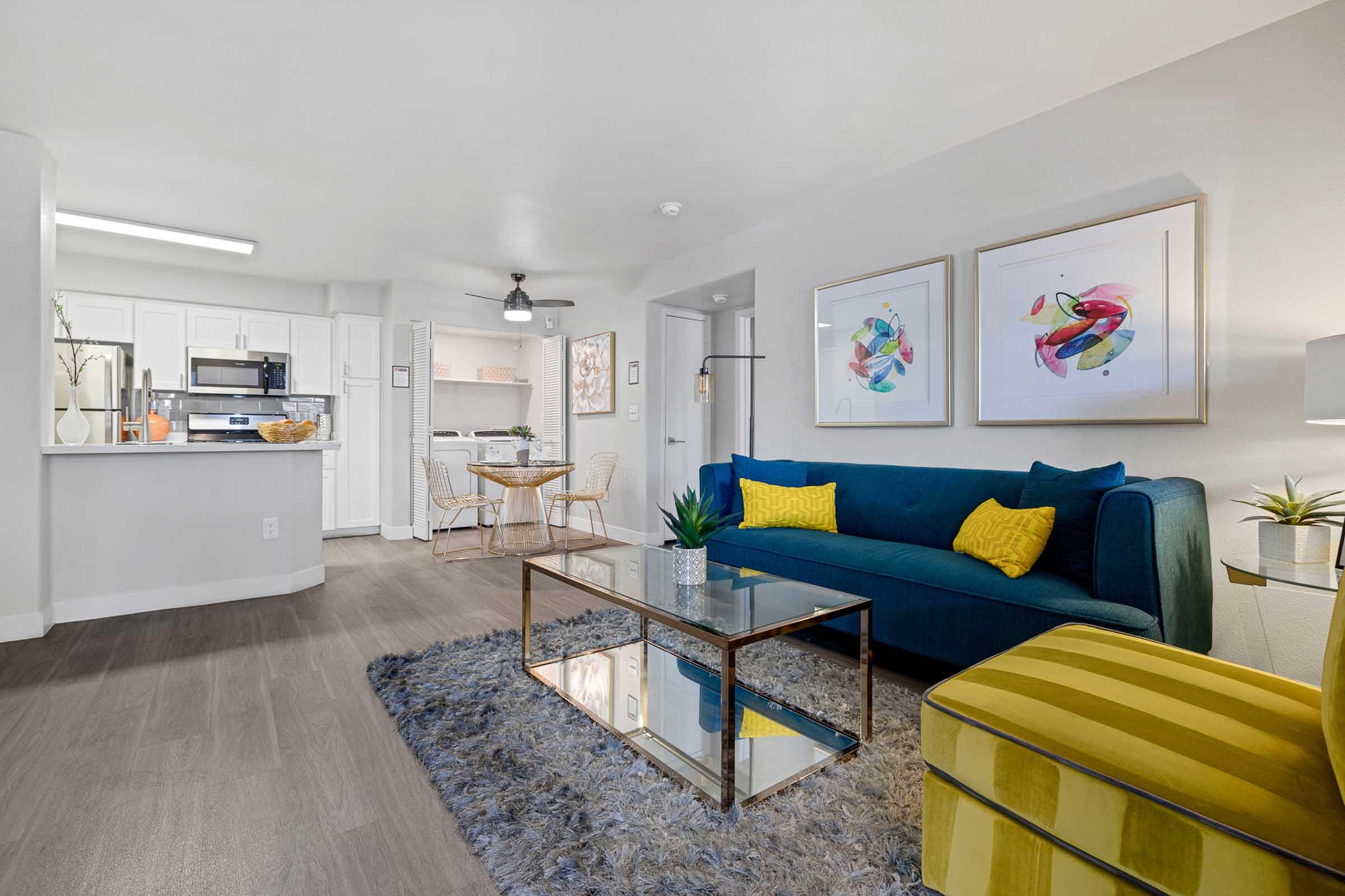 A cozy living room with a blue sofa adorned with yellow cushions, a glass coffee table on a fluffy rug, and two chairs around a small dining table. The kitchen is visible in the background, featuring modern appliances. Colorful abstract artwork decorates the wall, enhancing the contemporary aesthetic.