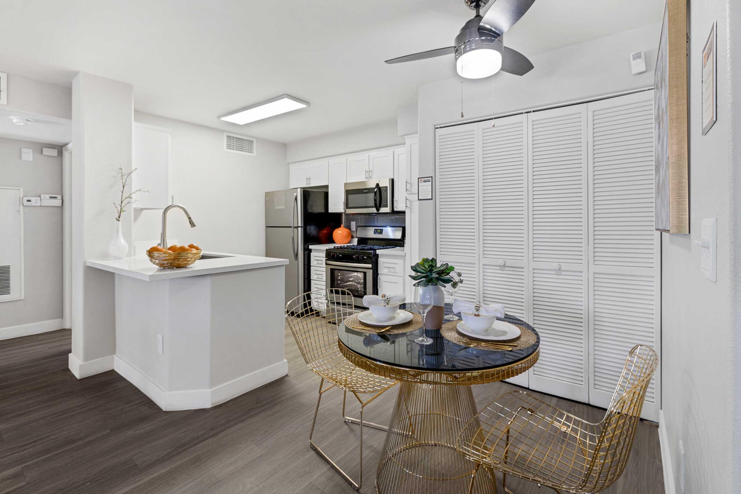 Modern kitchen and dining area featuring a round glass table with two place settings, golden wire chairs, and a small decorative bowl. The kitchen includes stainless steel appliances, a white countertop, and light cabinetry. Bright, neutral walls and a ceiling fan enhance the airy atmosphere.