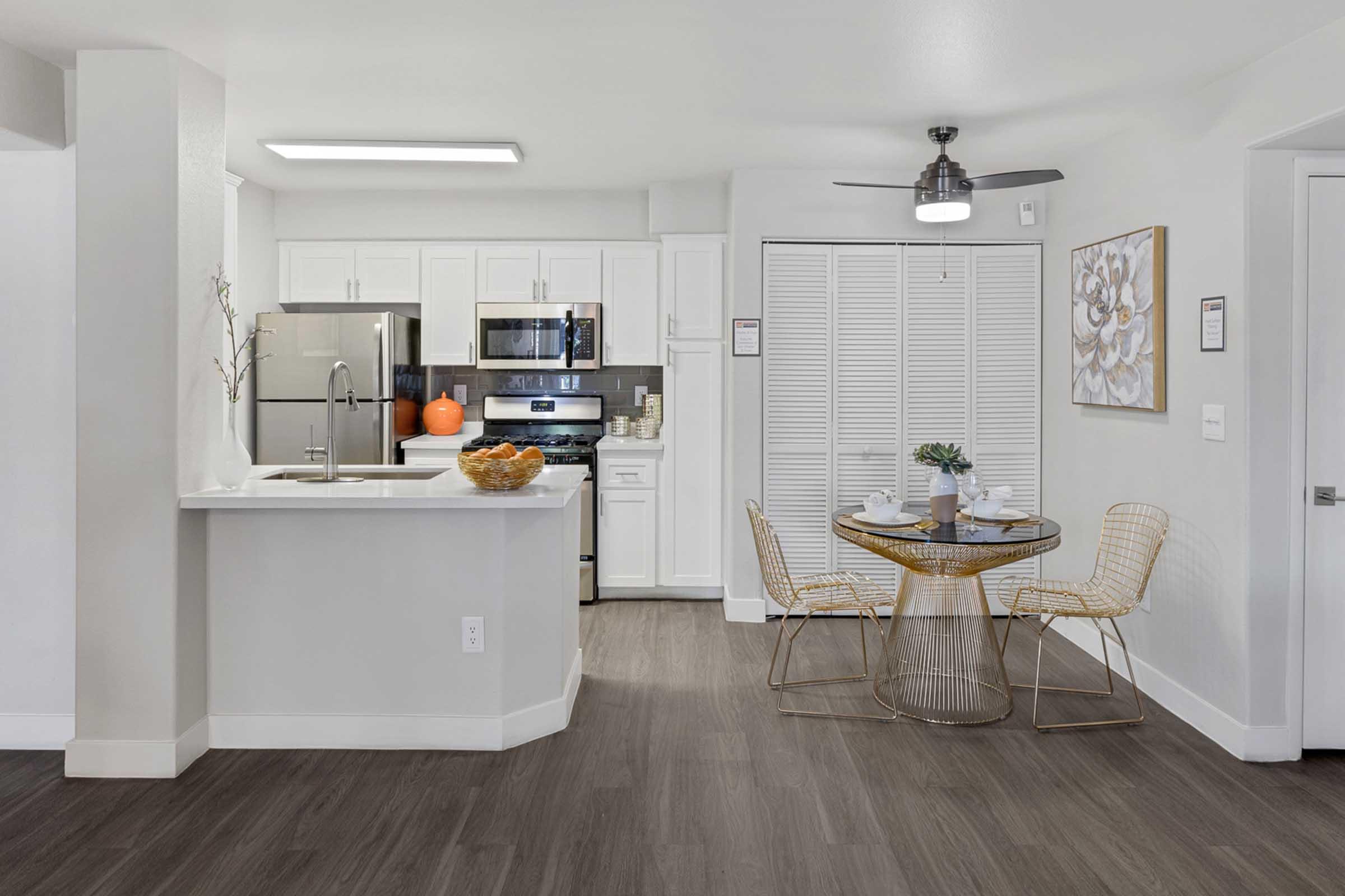 A modern kitchen area featuring white cabinets and stainless steel appliances, including a refrigerator and oven. A dining space with a round table and gold wire chairs is situated next to a window. The floor has a wood-like finish, and a decorative wall piece adds a touch of style.