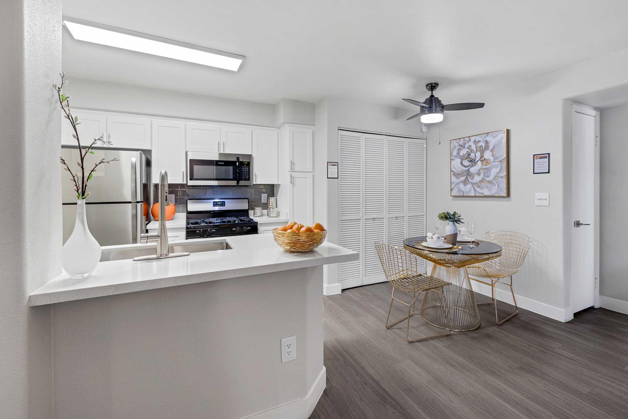 A modern kitchen featuring a white countertop, stainless steel appliances, and a breakfast nook with a round table and wire chairs. A bowl of oranges sits on the counter, and there's a ceiling fan above. The space has light-colored walls and wooden flooring, creating a bright and inviting atmosphere.