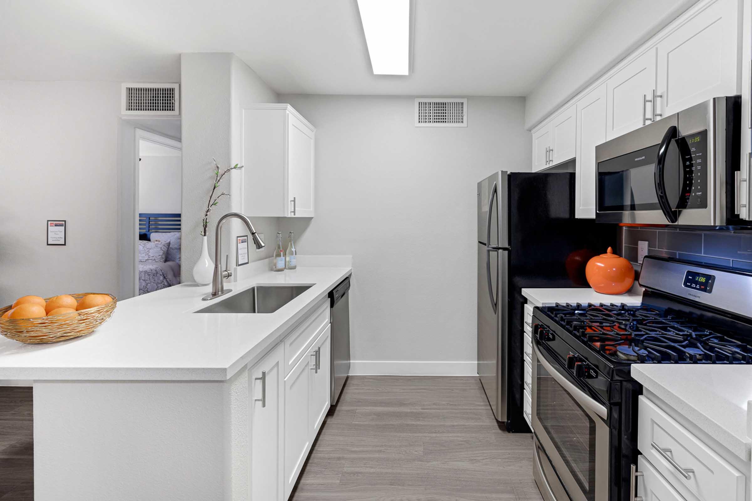 Modern kitchen featuring white cabinets, a stainless steel sink, a microwave, and a gas stove. A black refrigerator stands beside the stove. A bowl of oranges is placed on the counter, and there is an orange decorative pumpkin. The space is well-lit with a bright ceiling light.