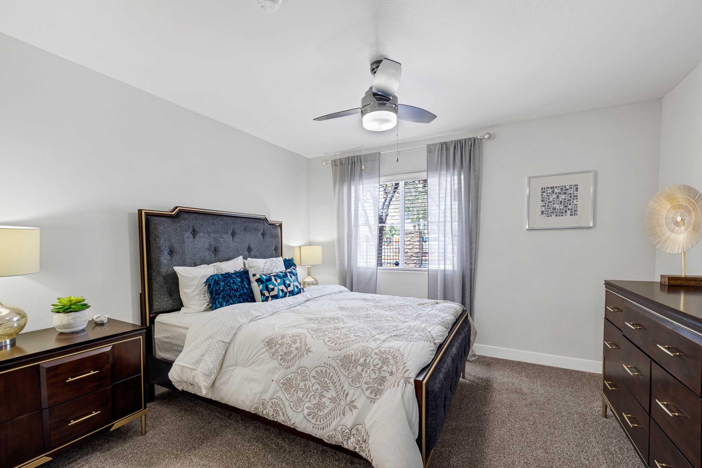 A stylish bedroom featuring a dark upholstered bed with decorative pillows, a nightstand with a lamp, and a dresser. Light gray walls complement the warm tones of the furniture. A ceiling fan hangs above, and a window with sheer curtains lets in natural light, creating a cozy and inviting atmosphere.