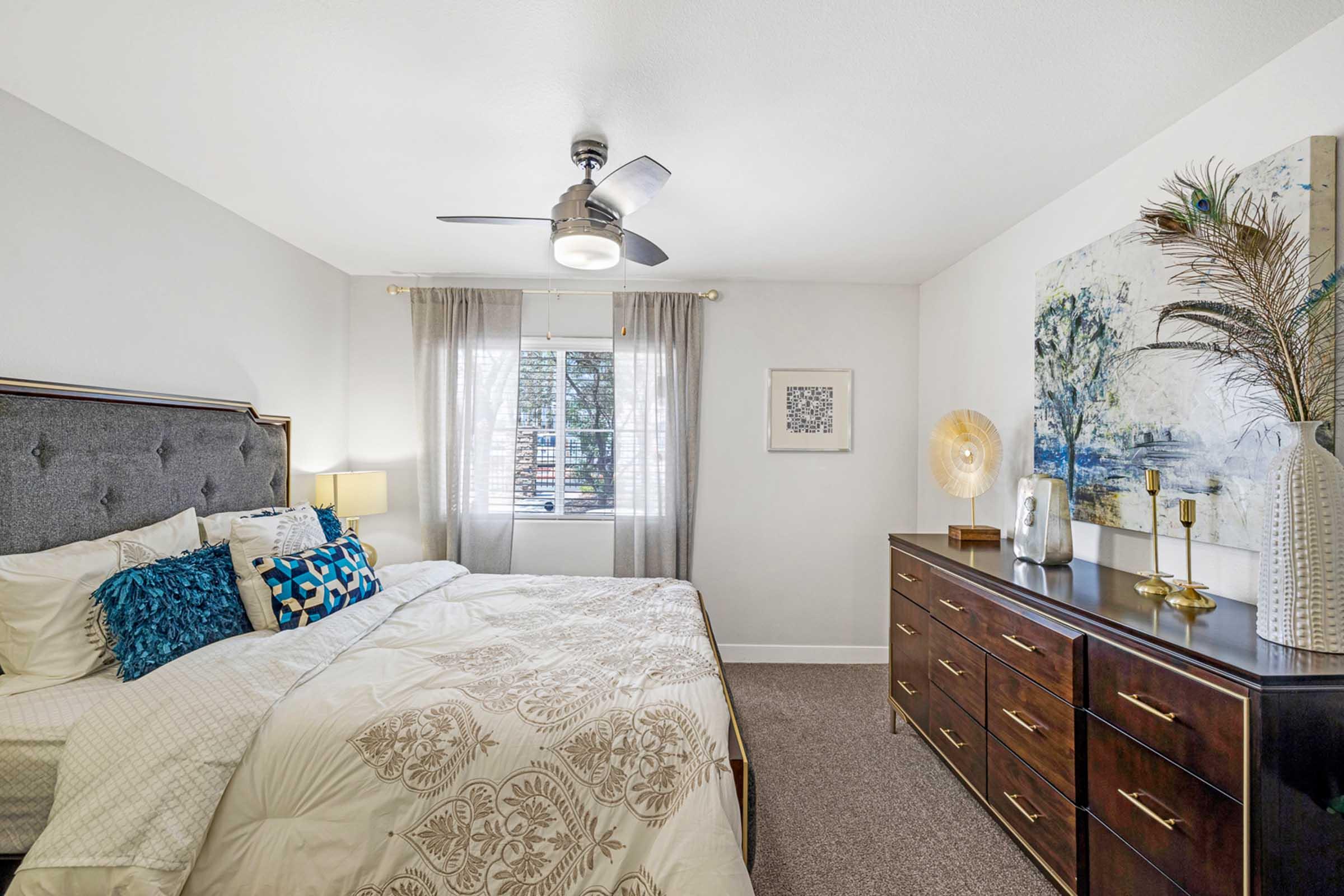 A cozy bedroom featuring a large bed with decorative pillows, a stylish gray headboard, and a soft beige bedding set. There’s a modern ceiling fan, natural light streaming through sheer curtains, and a dark wooden dresser adorned with decorative items and artwork on the wall.