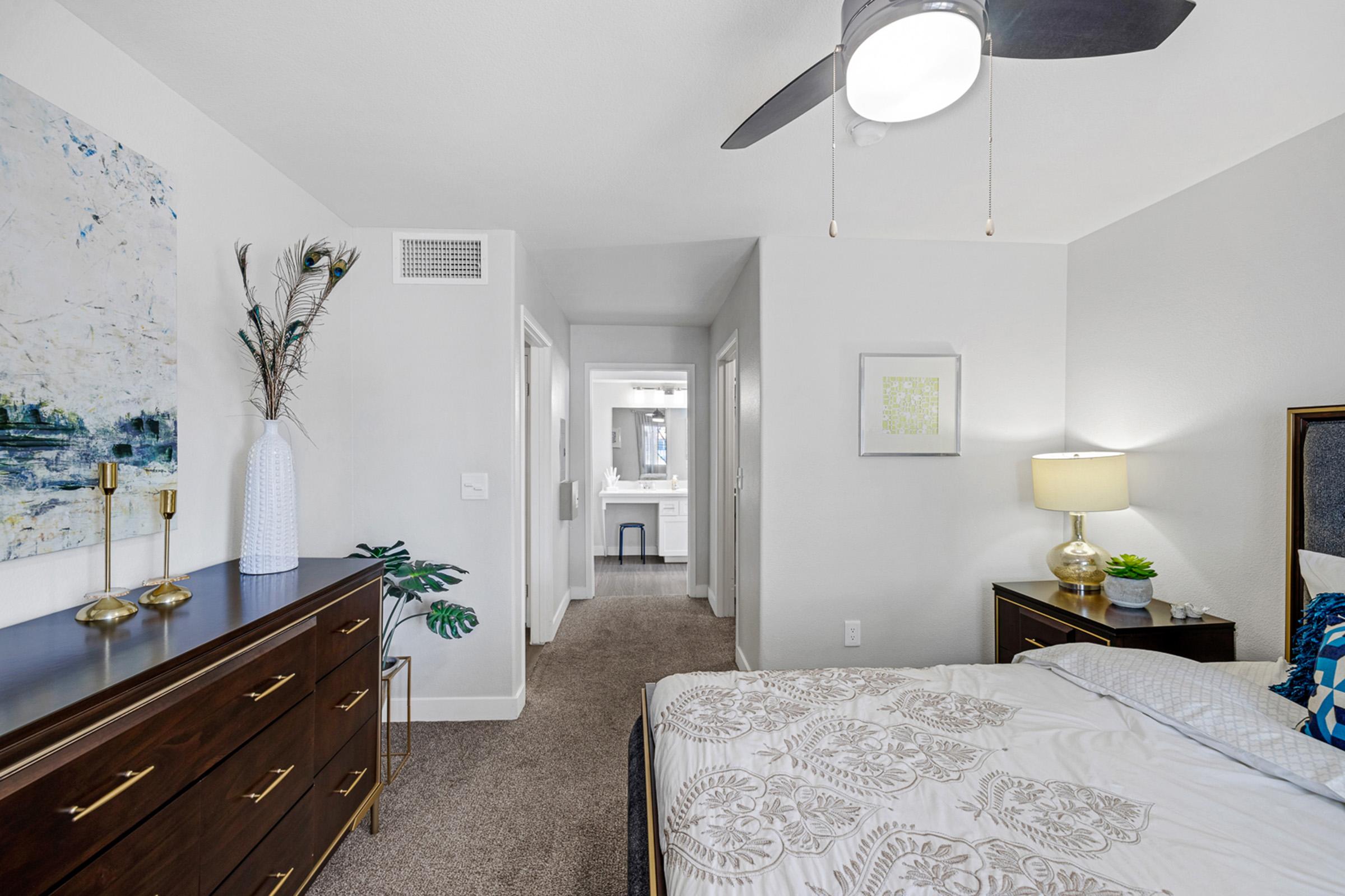 A modern bedroom featuring a comfortable bed with a decorative white and gray bedspread, a wooden dresser, and a bedside lamp. The walls are painted light gray, and a large piece of artwork is displayed. A doorway leads to a bathroom, with a cozy carpet and plants adding a touch of elegance.
