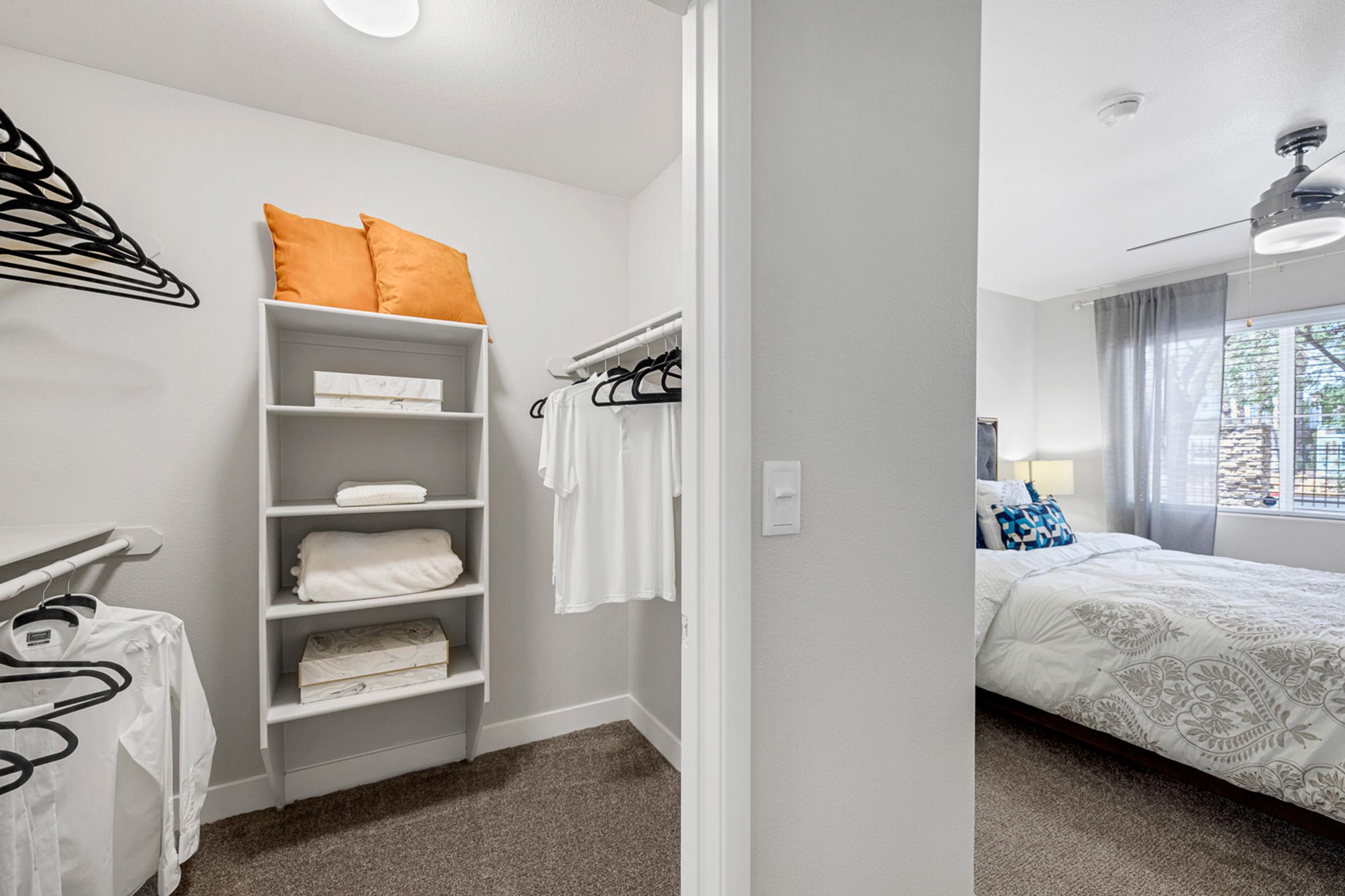 A well-organized closet featuring a shelf with neatly stacked towels and several hanging garments. Visible in the background is a cozy bedroom with a bed adorned in white and blue bedding, a window with sheer curtains, and a ceiling fan. The overall space is bright and inviting.
