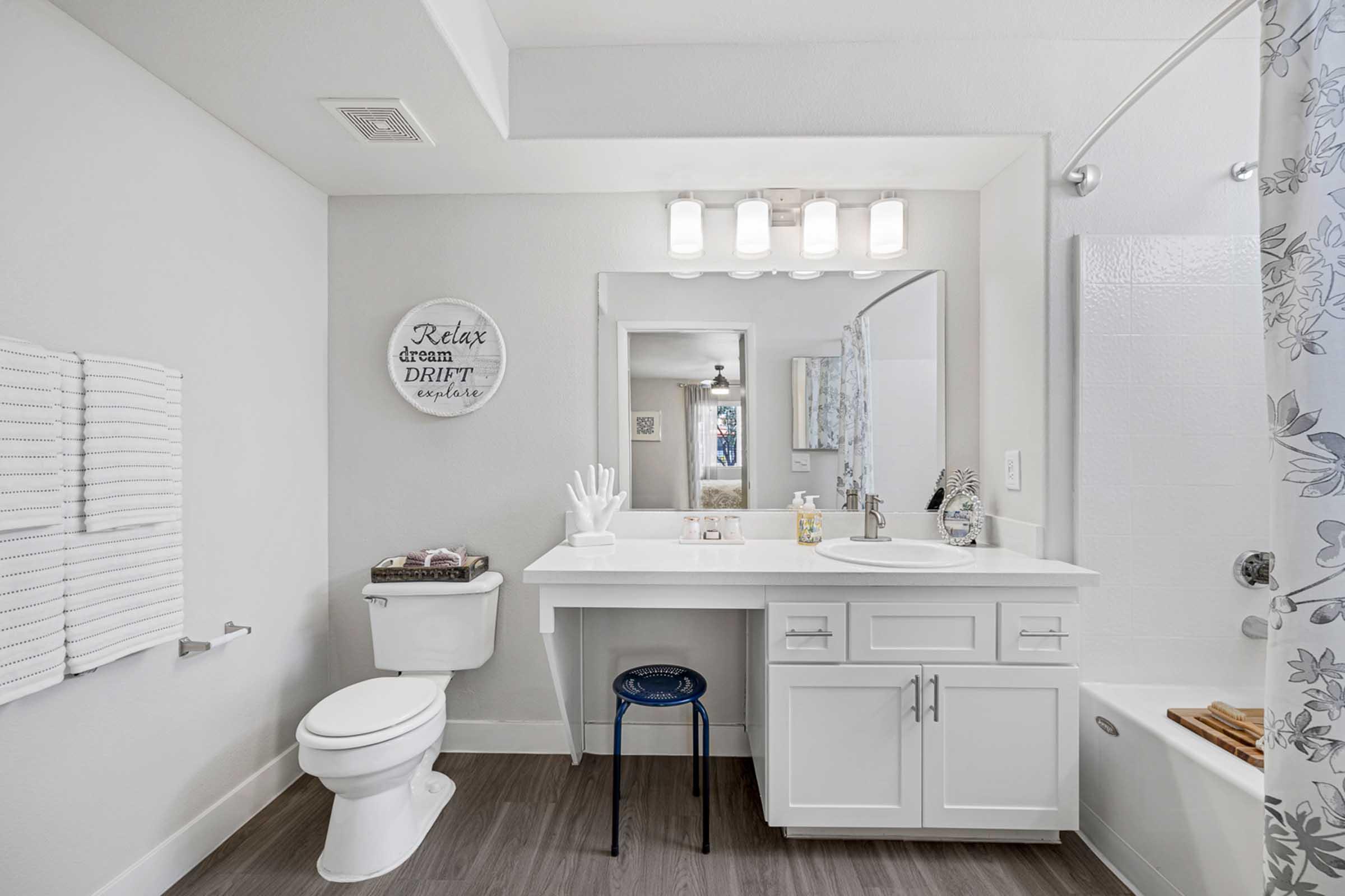 A modern bathroom featuring a white vanity with a large mirror, a sink, and decorative items. The space includes a toilet, striped towels on a rack, a blue stool, and a shower with a patterned curtain. Neutral walls and wood-like flooring create a clean, inviting atmosphere.