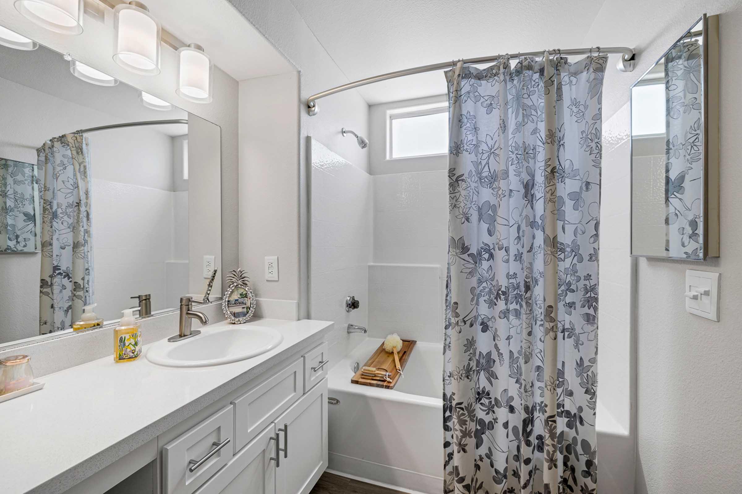 A modern bathroom featuring a white vanity with a round sink, a shower-tub combo with a floral curtain, a large mirror with lights above, and decorative items on the counter. The walls are light-colored, and there is a small window allowing natural light.