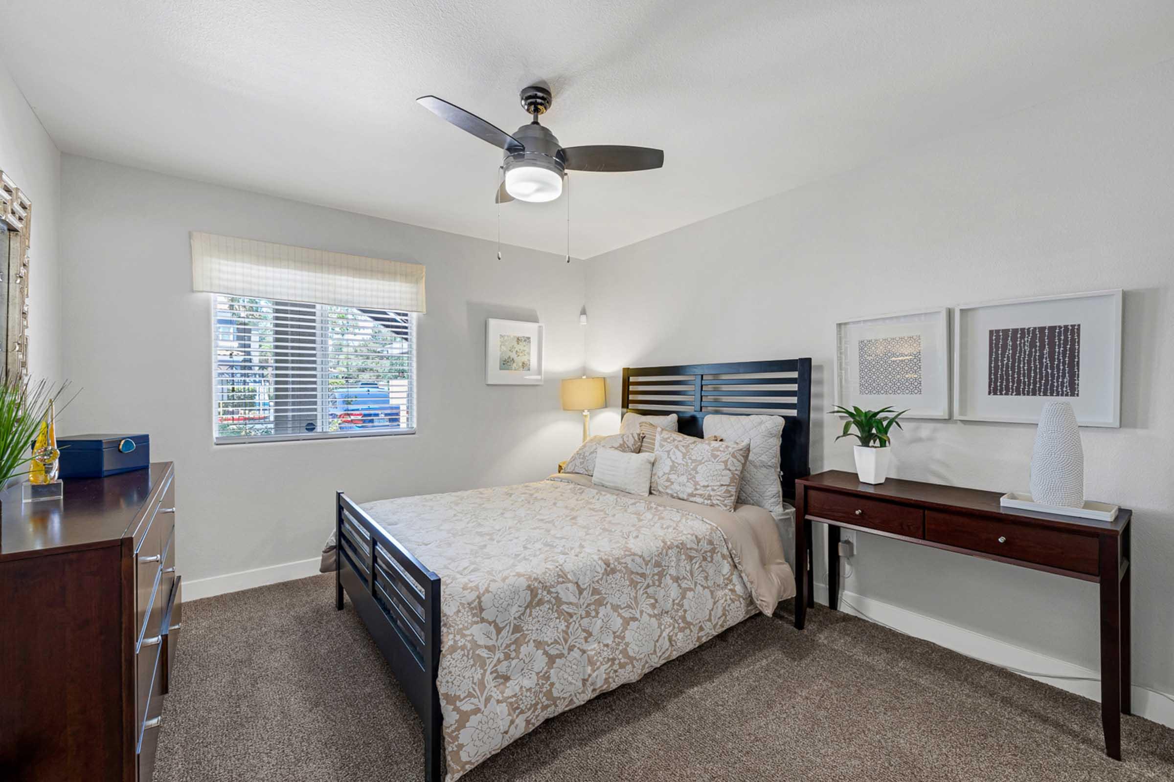 A well-decorated bedroom featuring a queen-sized bed with a patterned bedspread, a nightstand with a lamp, and a matching dresser. Natural light filters in through a window adorned with blinds. The walls are painted a soft color, enhancing the cozy ambiance. A plant and decorative items add a touch of warmth.
