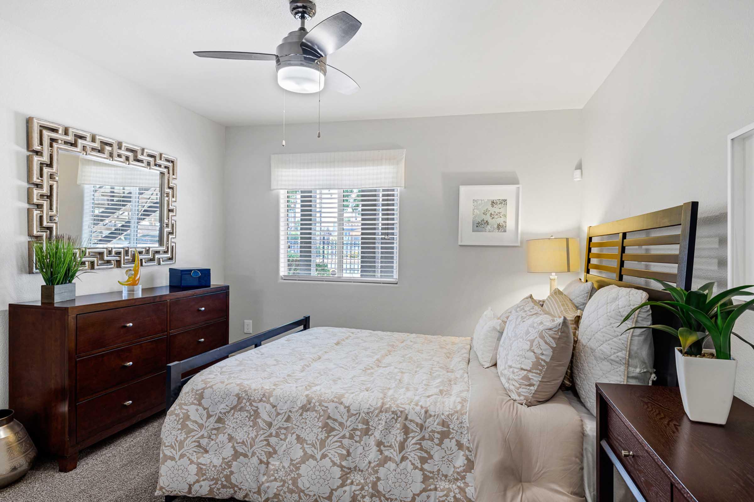 Cozy bedroom featuring a queen-sized bed with a floral quilt, a wooden headboard, and decorative pillows. A wooden dresser is positioned against one wall, accompanied by a mirror. Natural light filters through a window with blinds, while potted plants add a touch of greenery to the space. A ceiling fan provides ventilation.