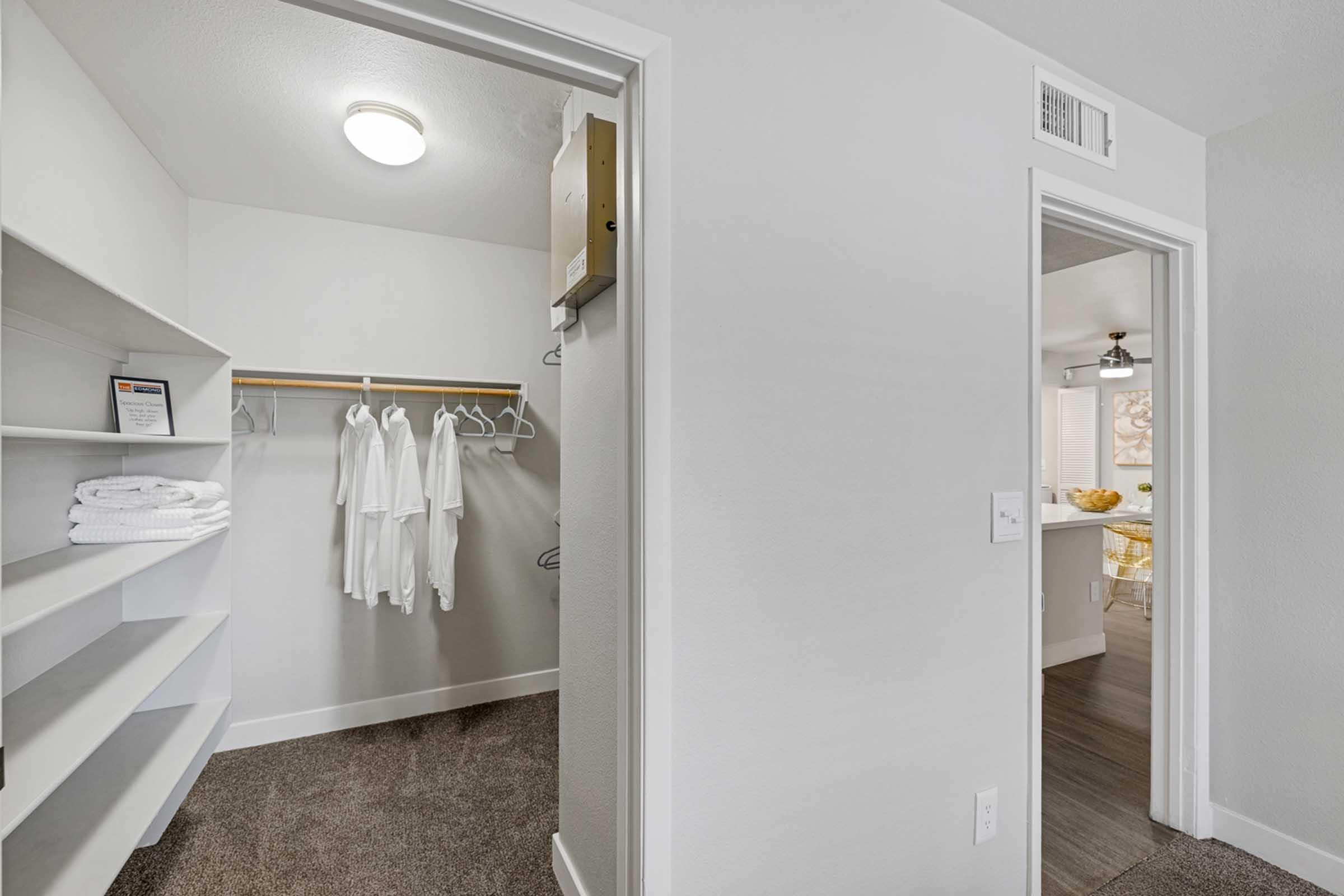 View of a small, minimalistic room with two doors. On the left, a closet features a shelf and hanging space with white clothing and towels. On the right, an open door leads to another room with visible natural light and a glimpse of a dining area. The walls are painted light colors, and the flooring is dark.
