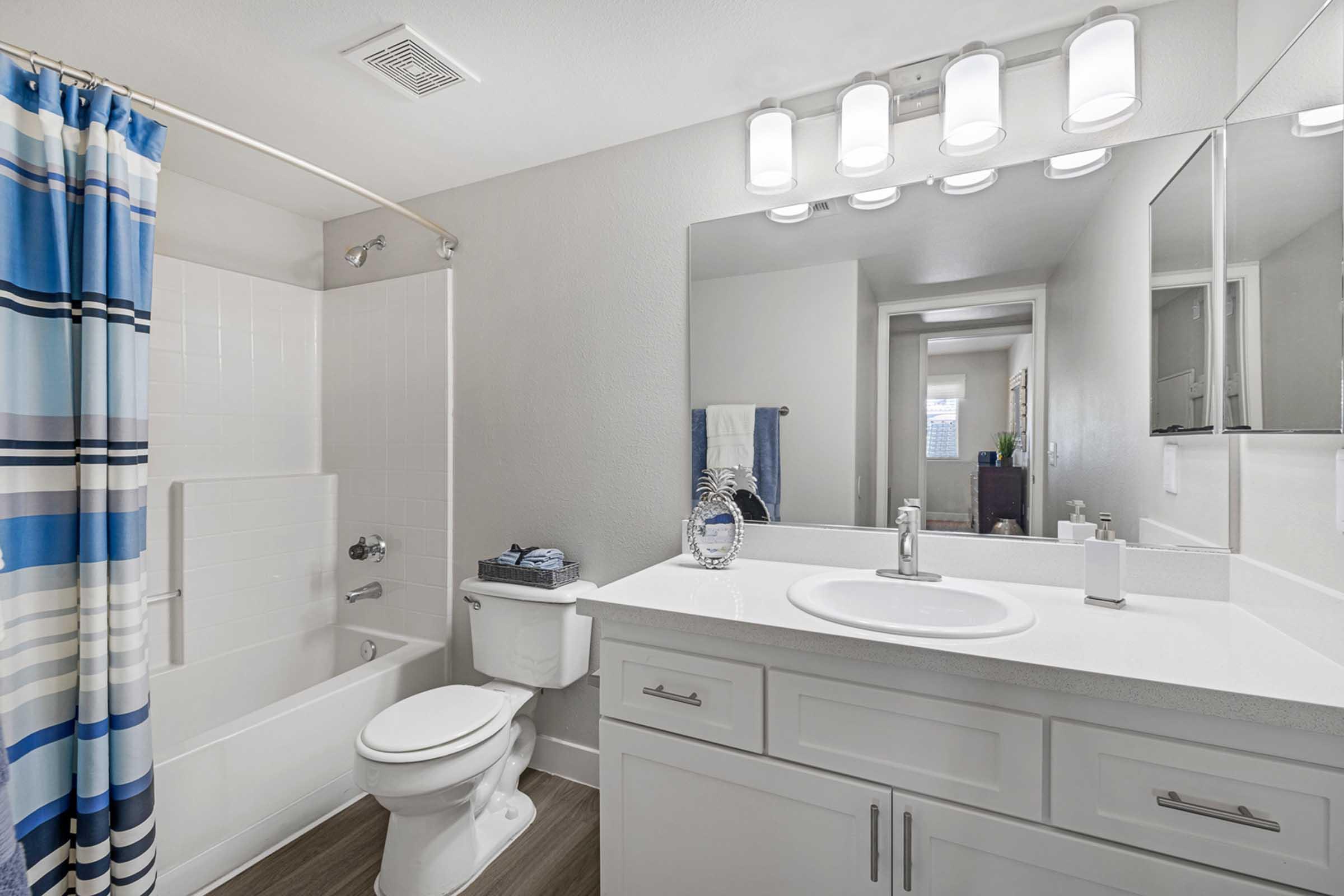 A modern bathroom featuring a white bathtub and shower combo, a white vanity with a sink, and a large mirror. The walls are painted a light color, and there's a striped shower curtain in blue and white. Soft lighting is provided by a series of overhead fixtures, enhancing the clean and bright ambiance.