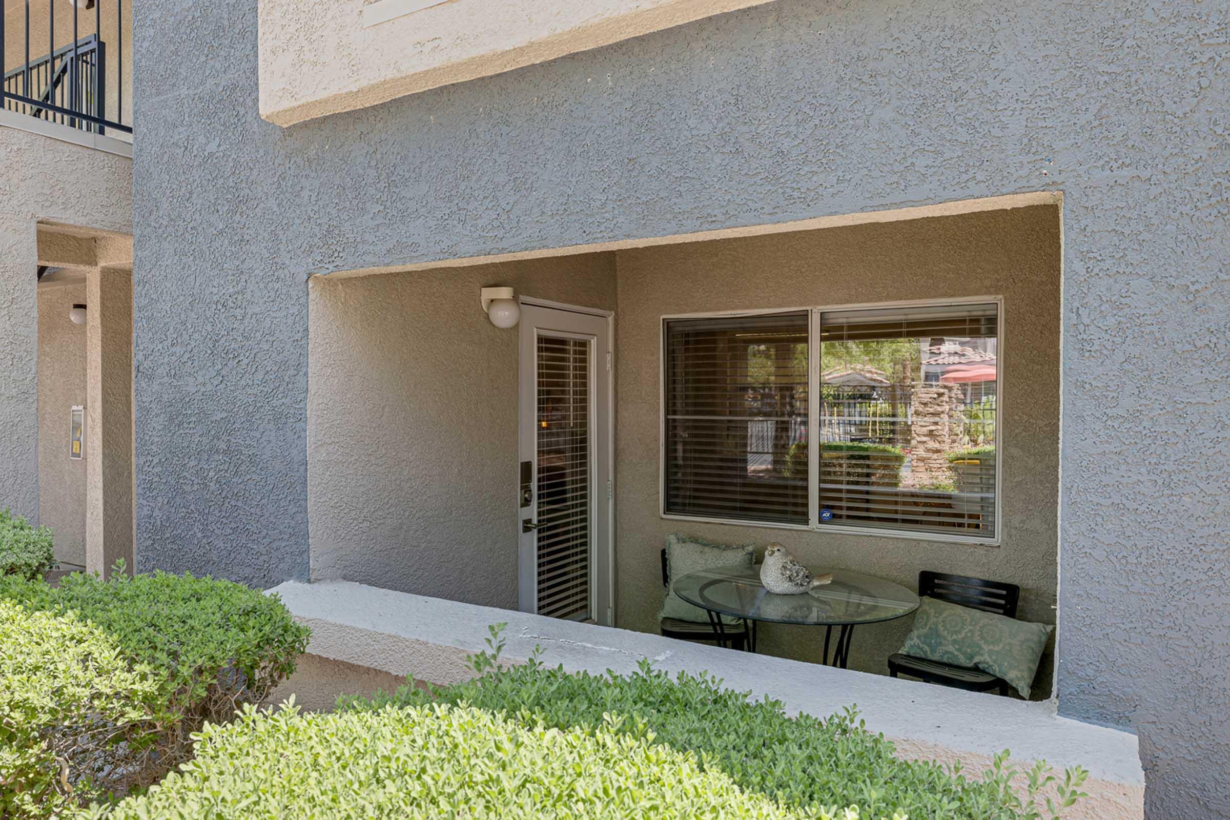 A small outdoor patio area of an apartment featuring a round glass table with two black chairs, surrounded by low green shrubs. A decorative item sits on the table, and a door leads into the apartment. The view shows a glimpse of the community outside.