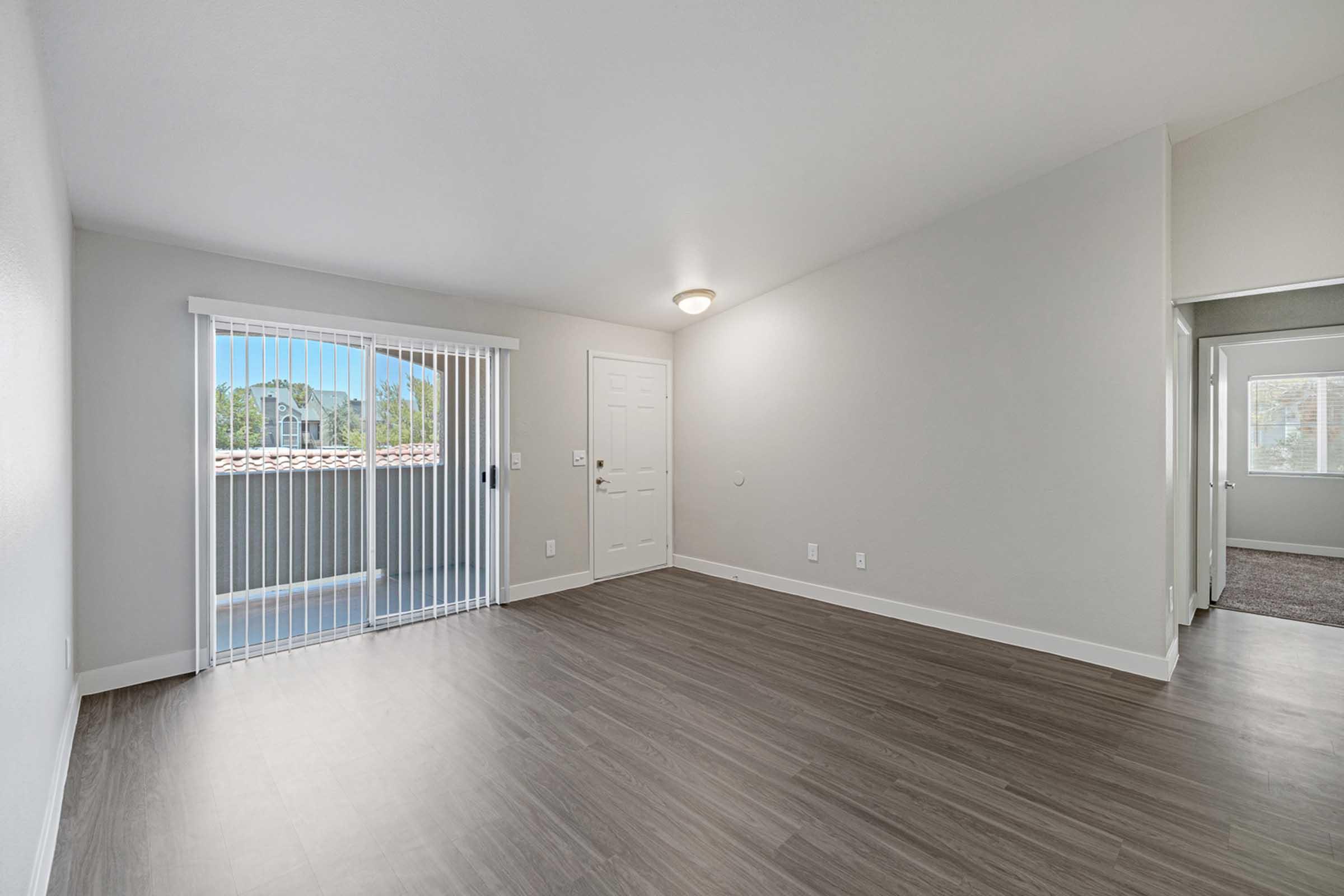 A bright and spacious living room with light-colored walls and laminate flooring. There is a sliding glass door leading to a balcony, letting in natural light. A ceiling light fixture is visible, and there is an open doorway leading to another room. The overall atmosphere is modern and inviting.