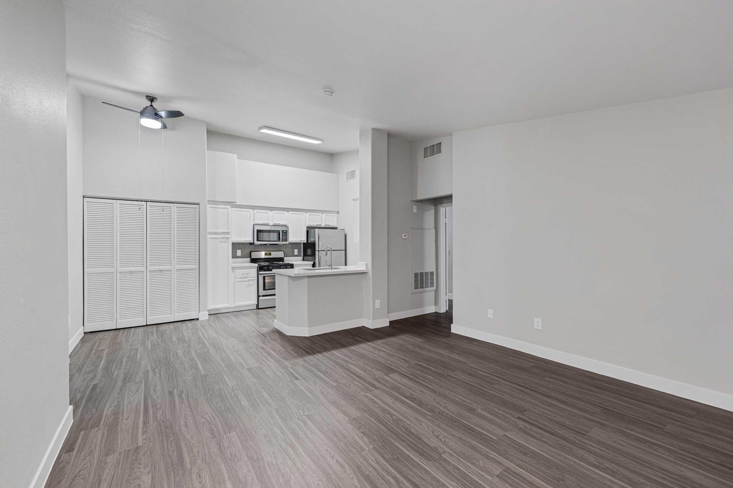 Modern, open-concept interior of an apartment featuring a kitchen with stainless steel appliances, a center island, and a spacious living area with wood-like flooring. White cabinetry and neutral wall colors create a bright, inviting atmosphere. A ceiling fan is visible in the room.