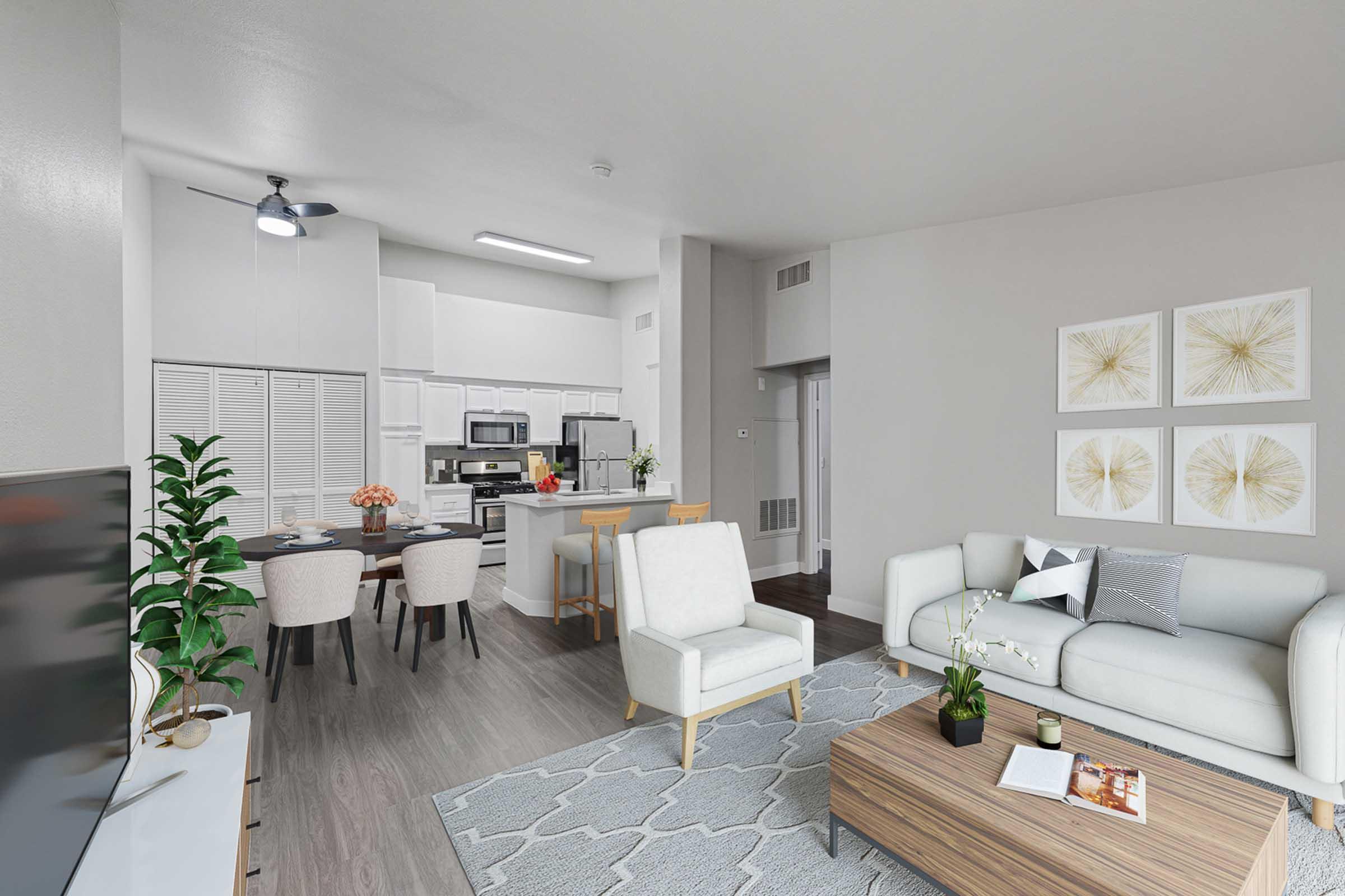 A modern living room and kitchen area featuring a white sofa, wooden coffee table, and patterned rug. In the background, there's a dining table set for four and a contemporary kitchen with white appliances. The walls are adorned with decorative art, and plants add a touch of greenery.
