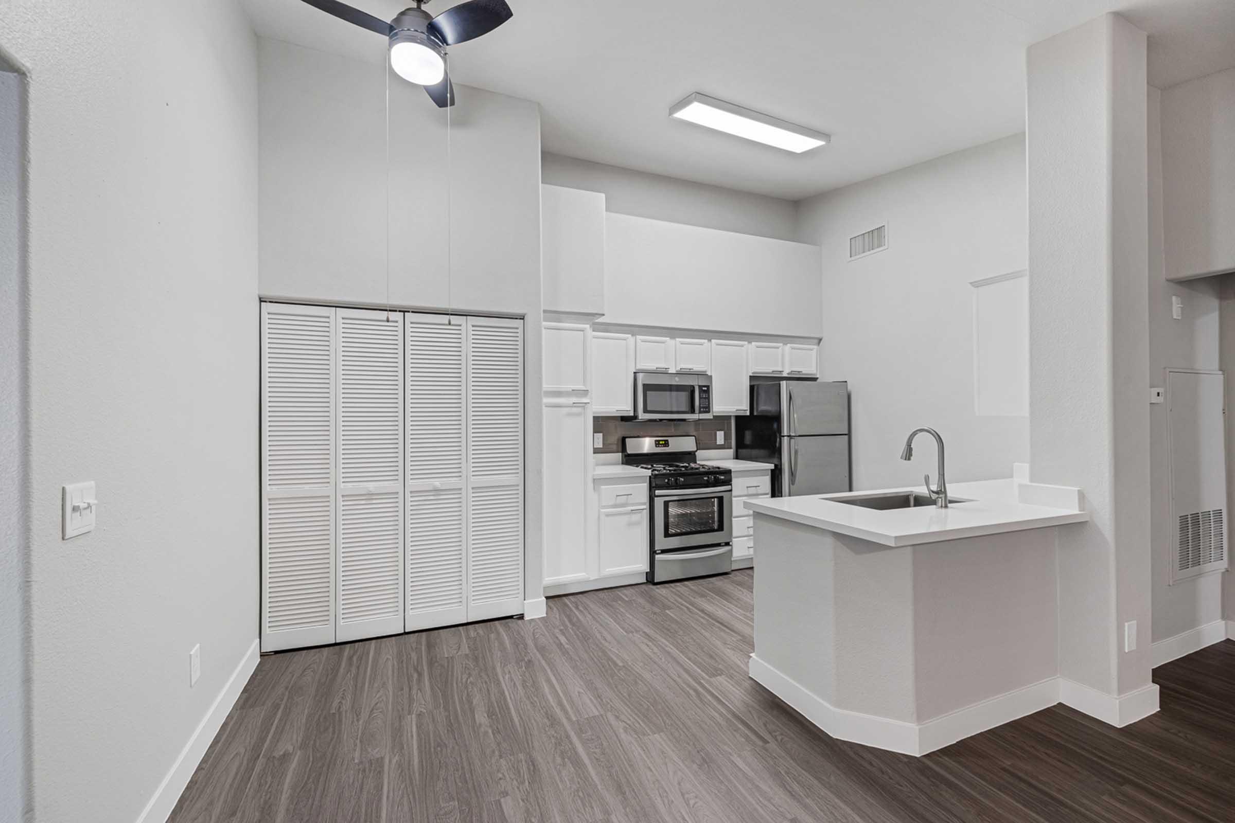 Modern kitchen interior featuring a white cabinetry, stainless steel appliances, and a kitchen island with a sink. The space has a light and airy feel with gray walls and dark wood flooring. Ceiling fan and overhead lighting add functionality. Ample storage with closed-door cabinets in the background.