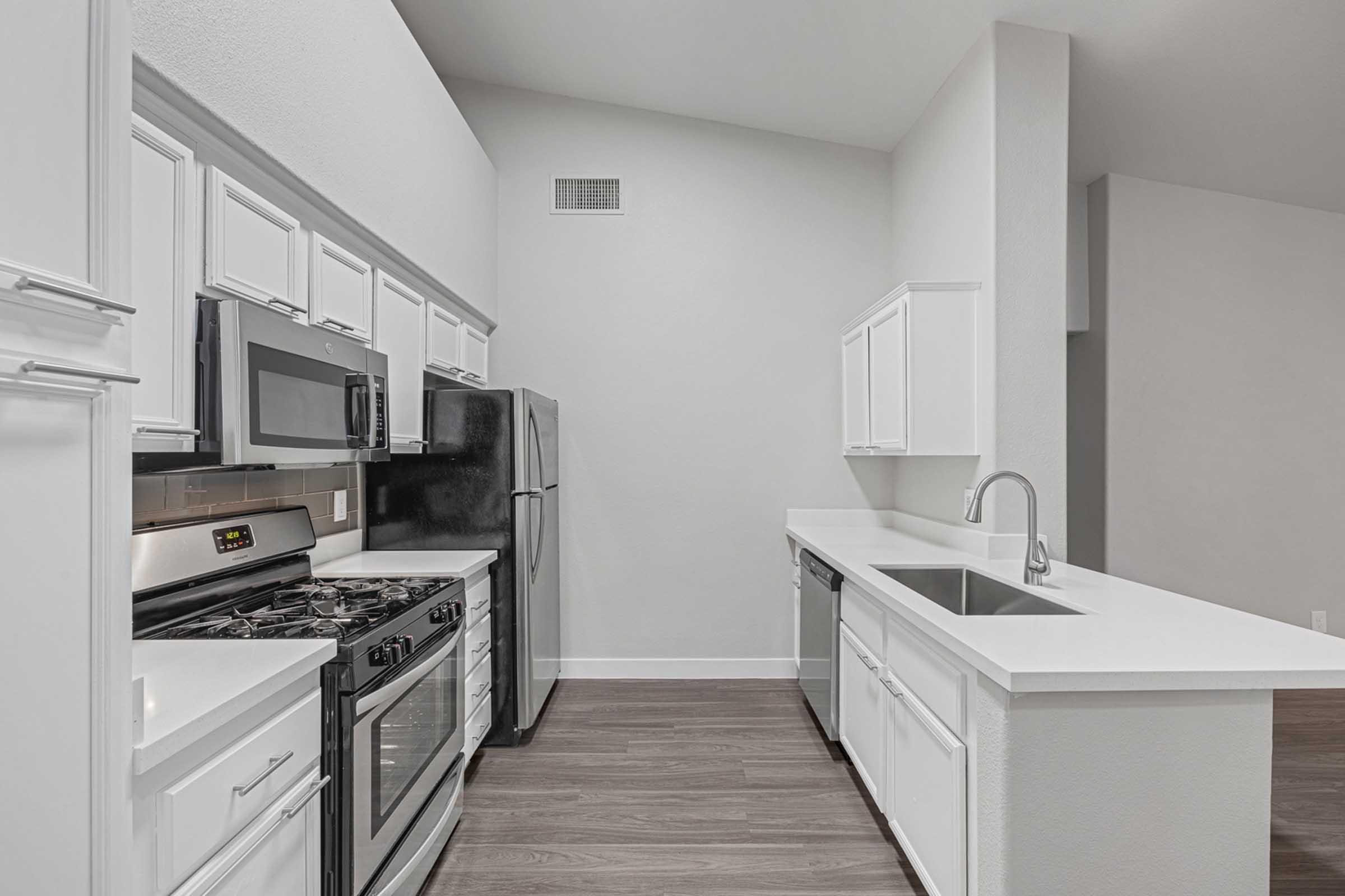 Modern kitchen featuring white cabinetry, stainless steel appliances including a microwave, gas stove, and refrigerator, alongside a sleek countertop and sink. The space has a light color scheme with clean lines and wooden flooring, providing a contemporary and inviting atmosphere.