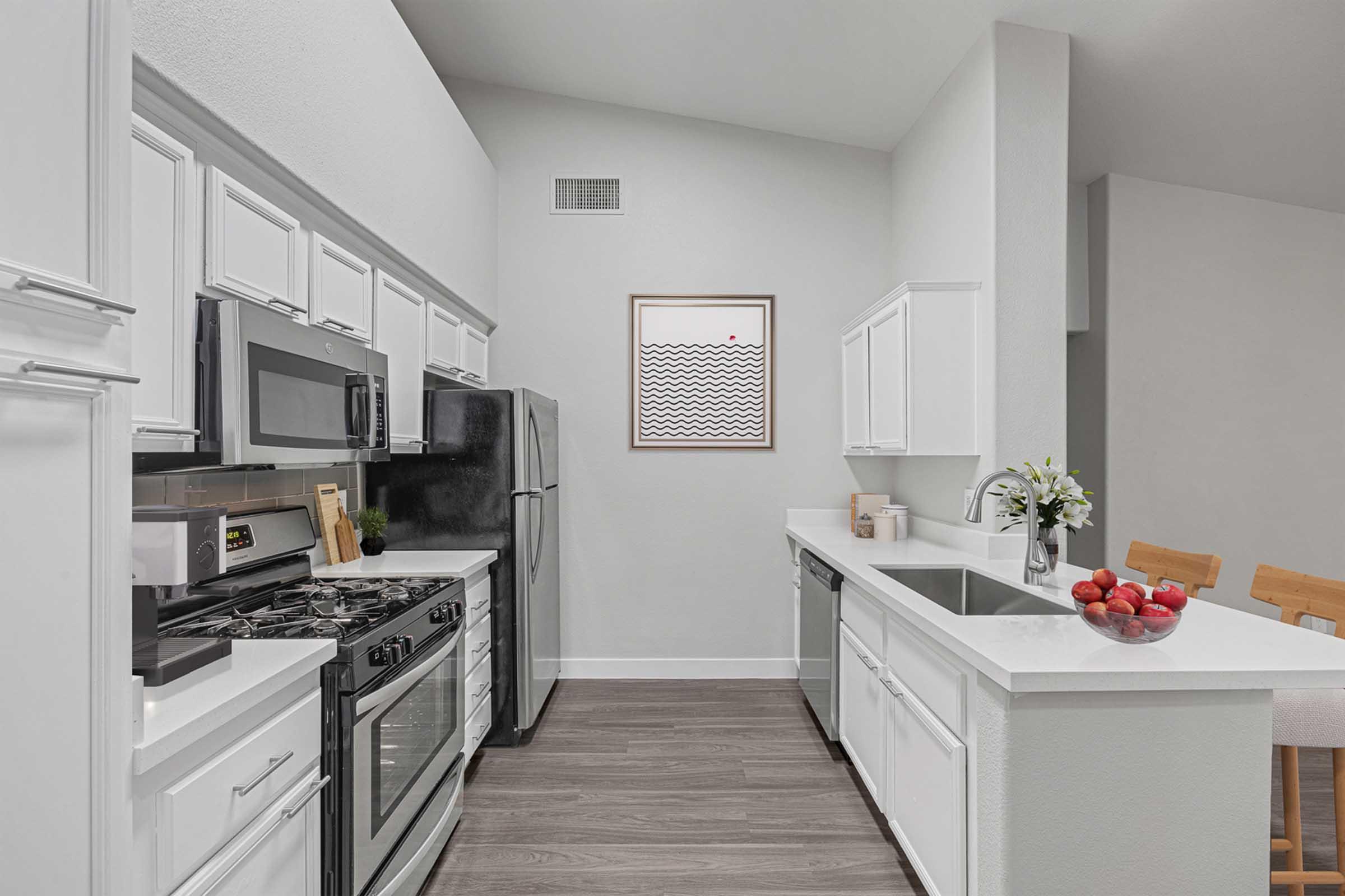 Modern kitchen featuring white cabinets, stainless steel appliances, and a gas stove. The countertop holds a bowl of red apples, and there's a framed abstract art piece on the wall. The space has a clean, minimalist design with natural light enhancing the ambiance.