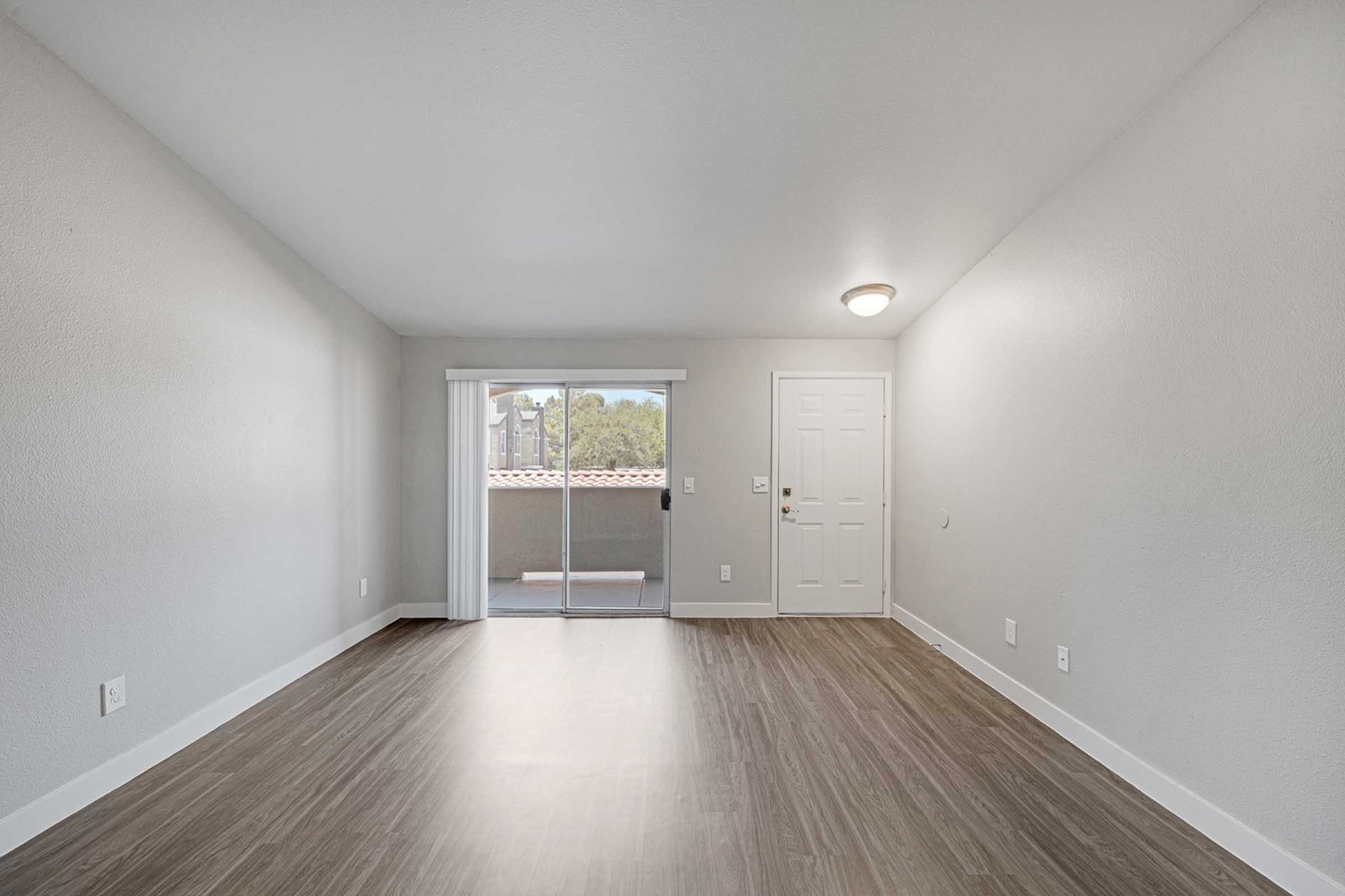 A spacious, empty room with light-colored walls and a wood-like floor. There is a sliding glass door leading to a balcony, allowing natural light to enter. The room features a single ceiling light and a front door on one wall. The overall aesthetic is modern and minimalistic.