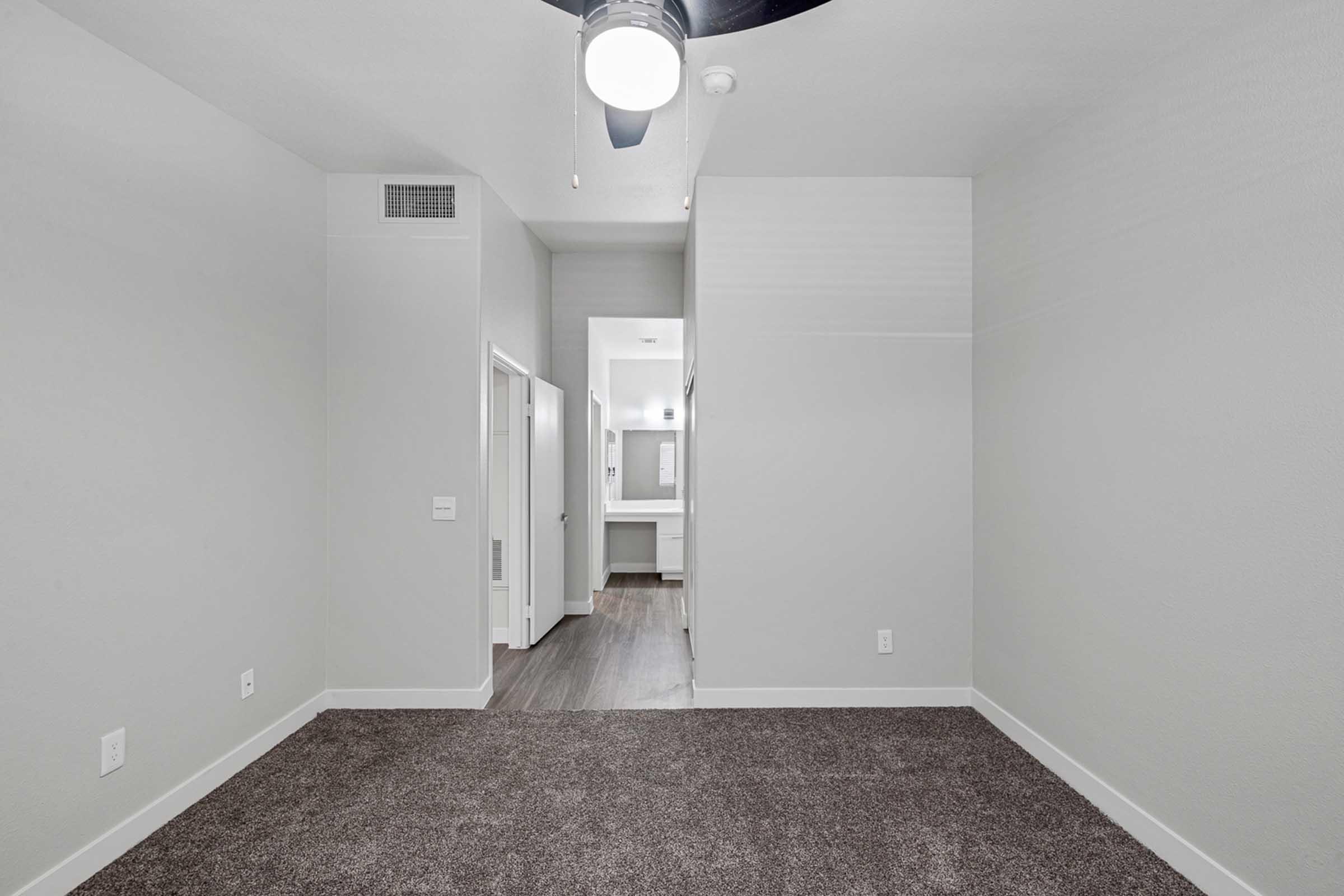 A spacious, brightly lit room with light gray walls and dark carpet flooring. A ceiling fan is mounted above, and there are doorways leading to other areas. The background features a hallway that opens to a well-lit area with a mirror and vanity, giving a sense of openness in the design.