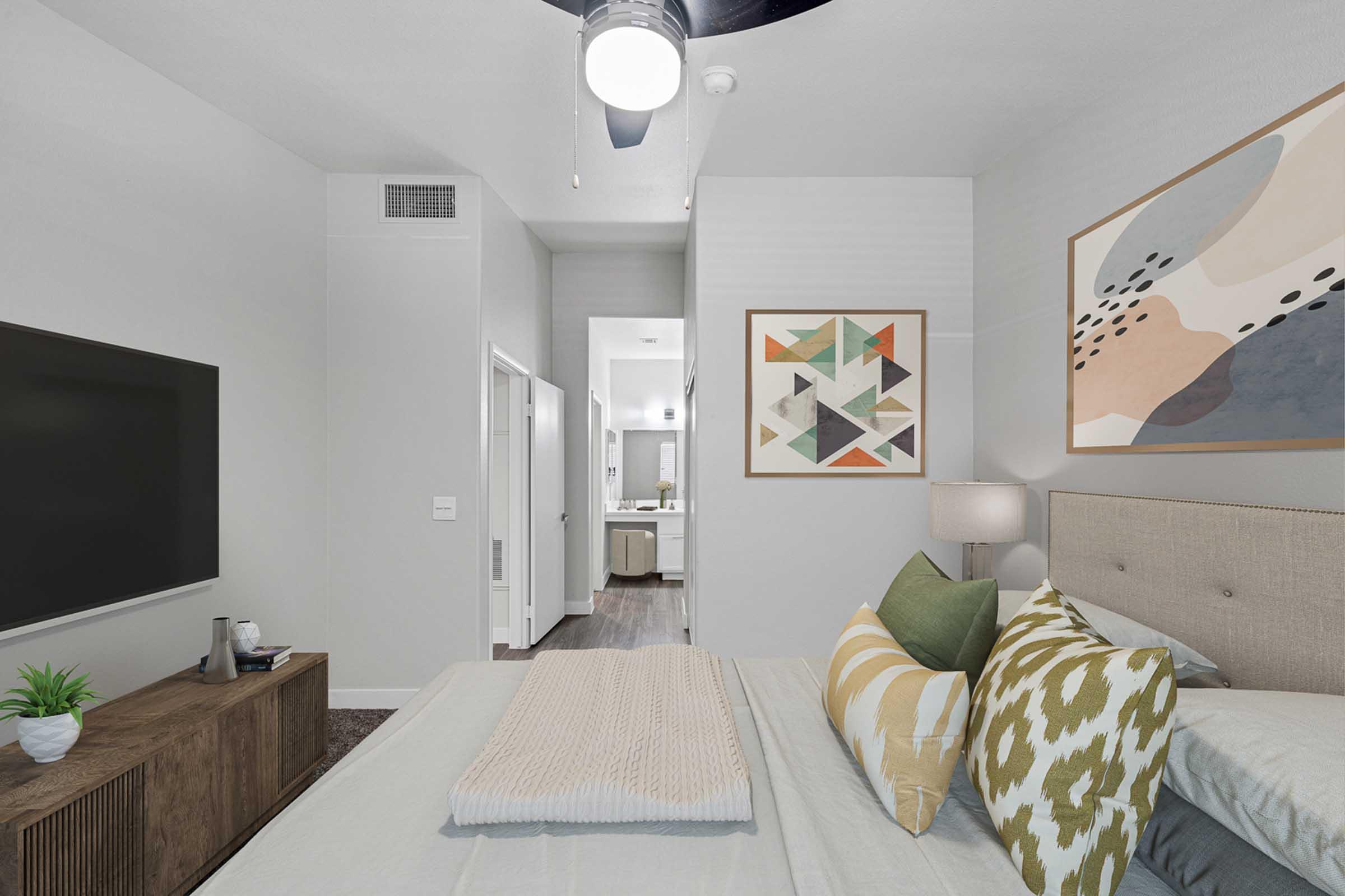A modern bedroom featuring a large bed with decorative pillows, a wall-mounted TV, and a bedside lamp. The walls are painted in light gray, and abstract art hangs above the bed. A doorway leads to a bathroom visible in the background, and a small plant is placed on the wooden dresser.