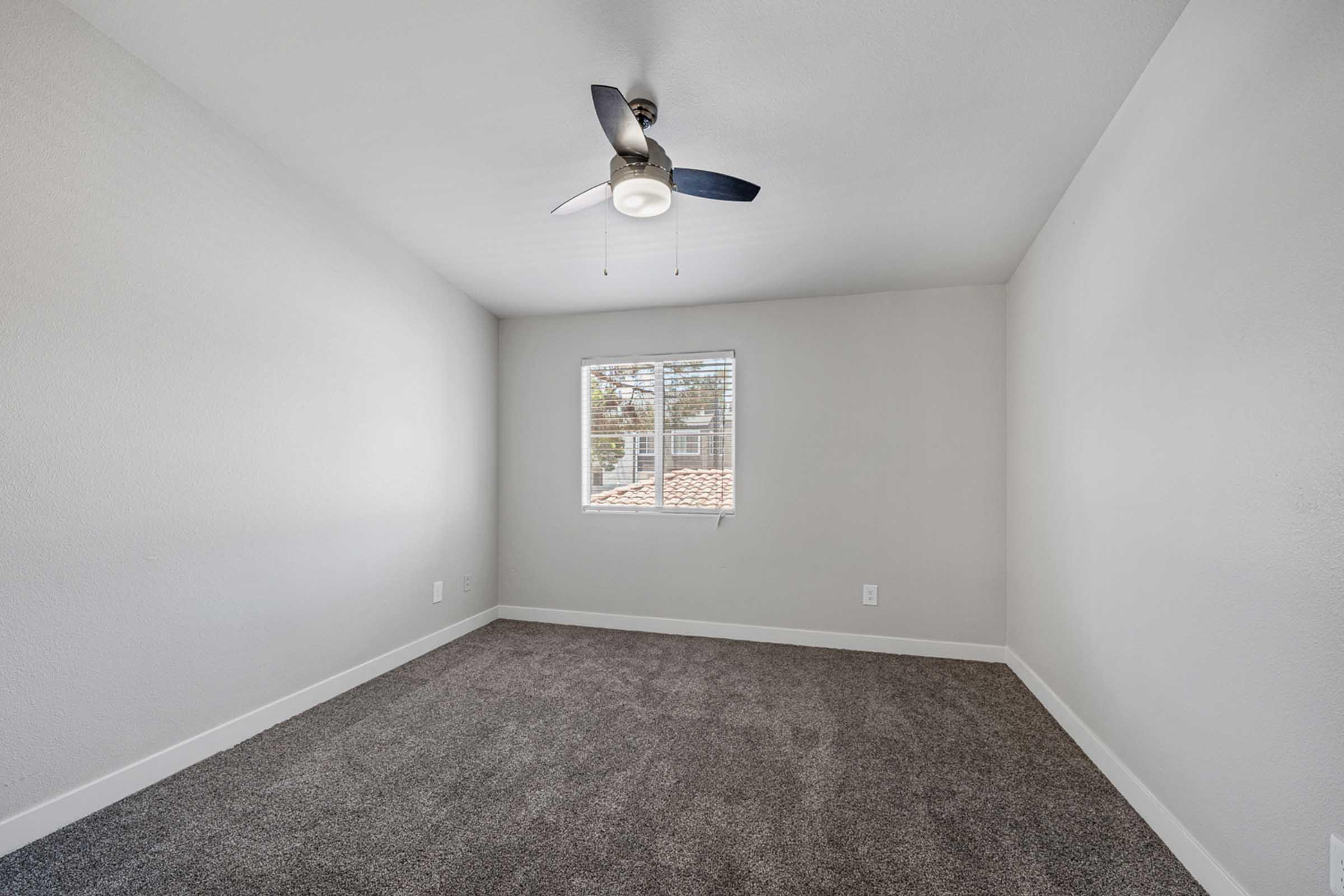 A bright, empty room with a vaulted ceiling and a ceiling fan. The walls are painted a soft gray, and there is light gray carpeting on the floor. A window with a view of a tiled roof allows natural light to enter, creating a spacious and airy atmosphere. No furniture is present in the room.