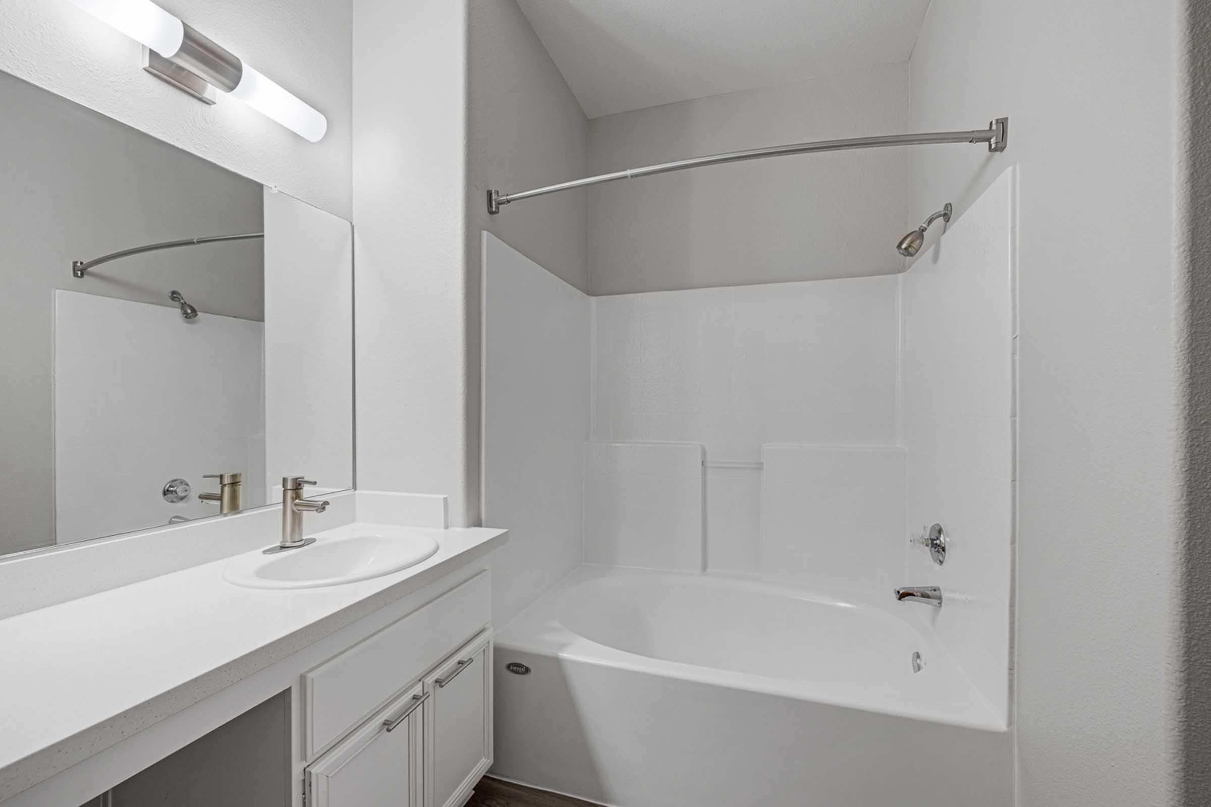 A modern bathroom featuring a white bathtub with a curved shower rod, a matching sink with a chrome faucet, and a large mirror. The walls are smooth and light-colored, contributing to a clean and bright atmosphere. The overall design emphasizes simplicity and functionality.