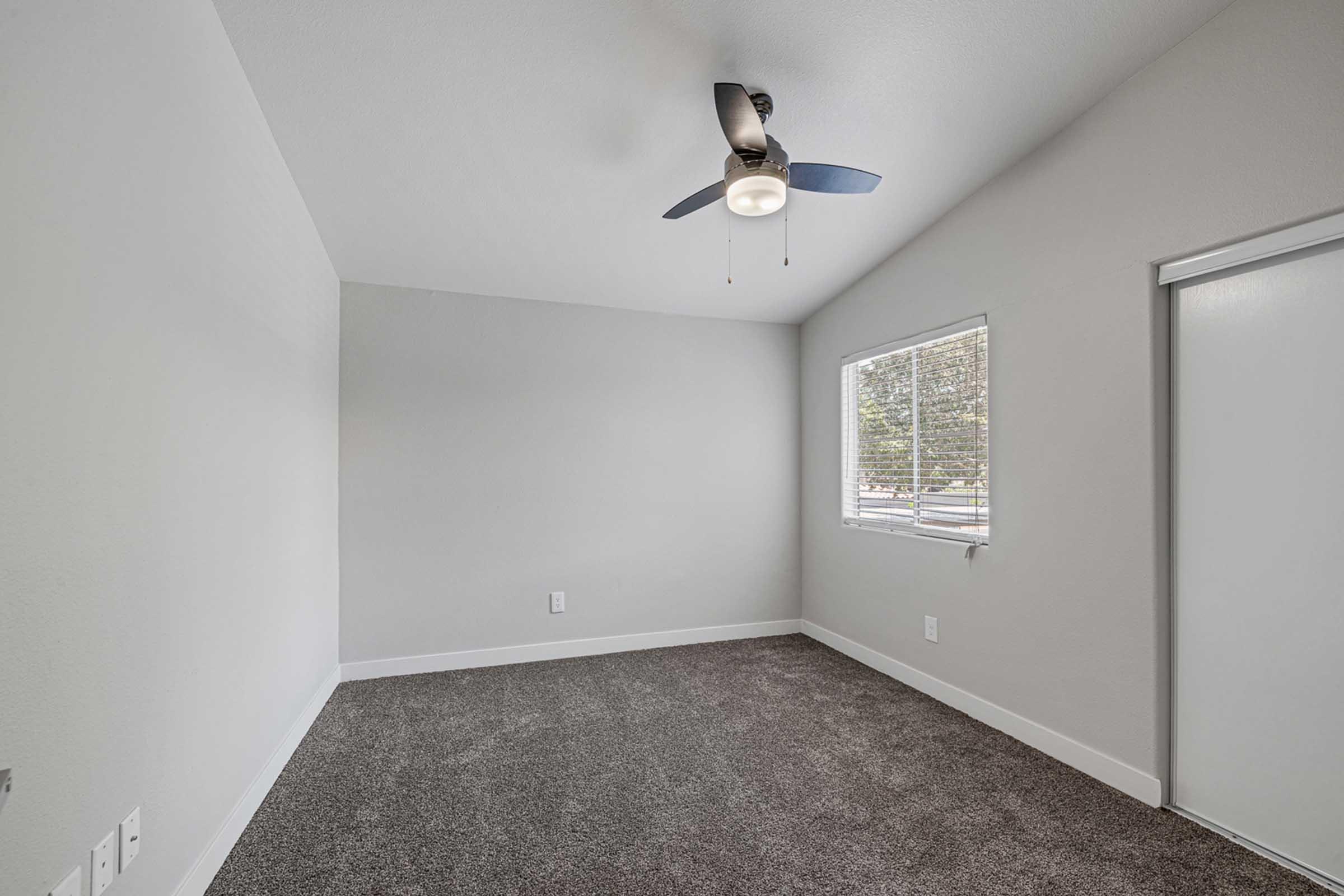 A clean, empty bedroom featuring light gray walls and a ceiling fan. It has a window allowing natural light and a door on the right. The floor is covered with a soft, dark gray carpet, creating a cozy atmosphere.