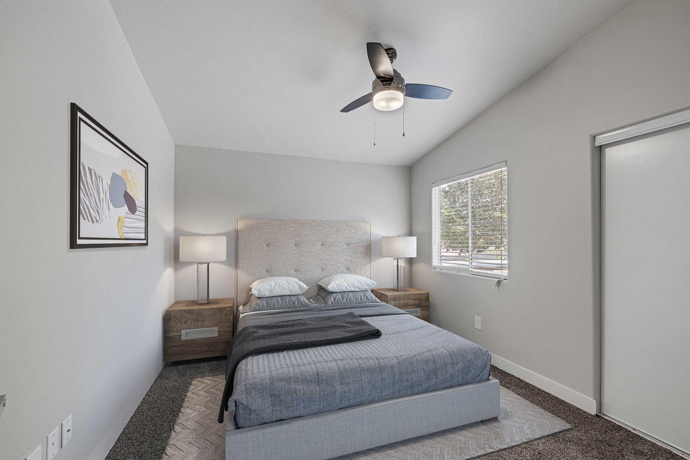 A modern bedroom featuring a light gray wall, a queen-sized bed with gray bedding, and two bedside lamps on wooden nightstands. A ceiling fan is installed above the bed, and a window with white blinds lets in natural light. A piece of artwork is hung on the wall, adding a decorative touch.