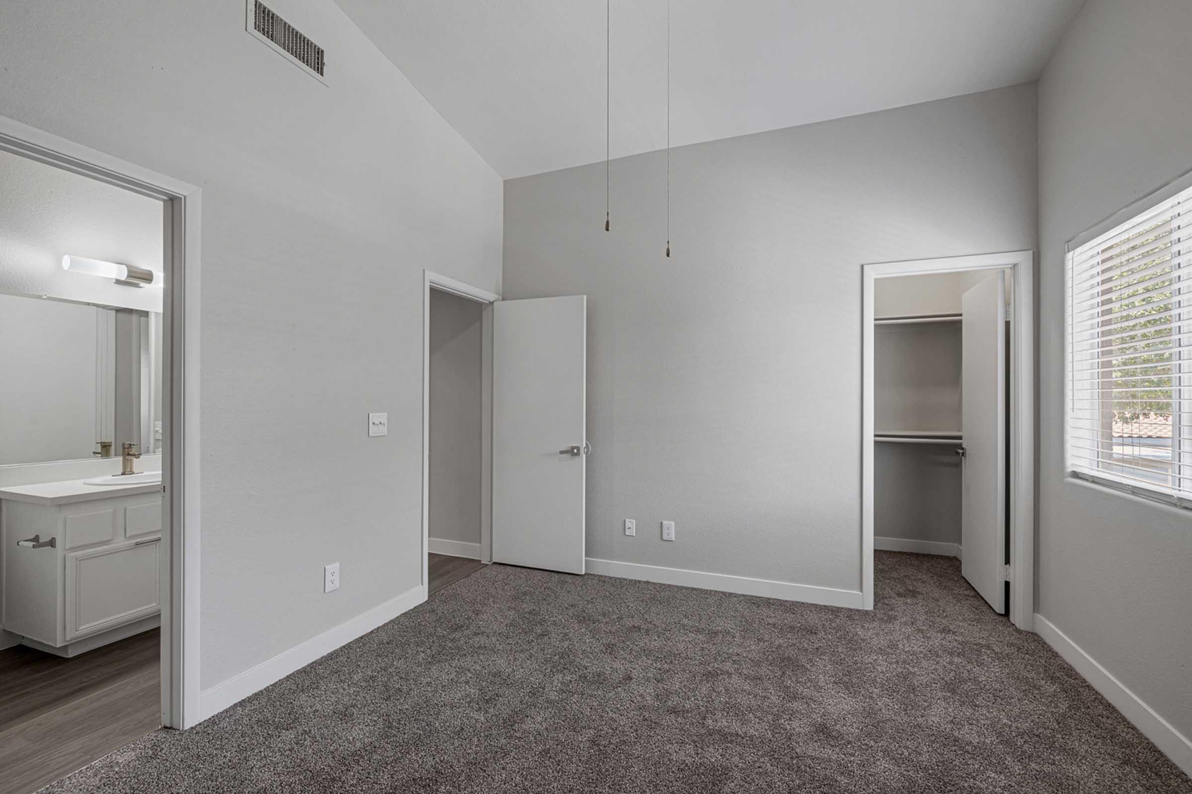 A well-lit, empty bedroom featuring light gray walls and carpet. A doorway leads to a closet on the right, while a bathroom is visible through an open door on the left. The room has two ceiling lights and a large window with blinds, allowing natural light to enter.