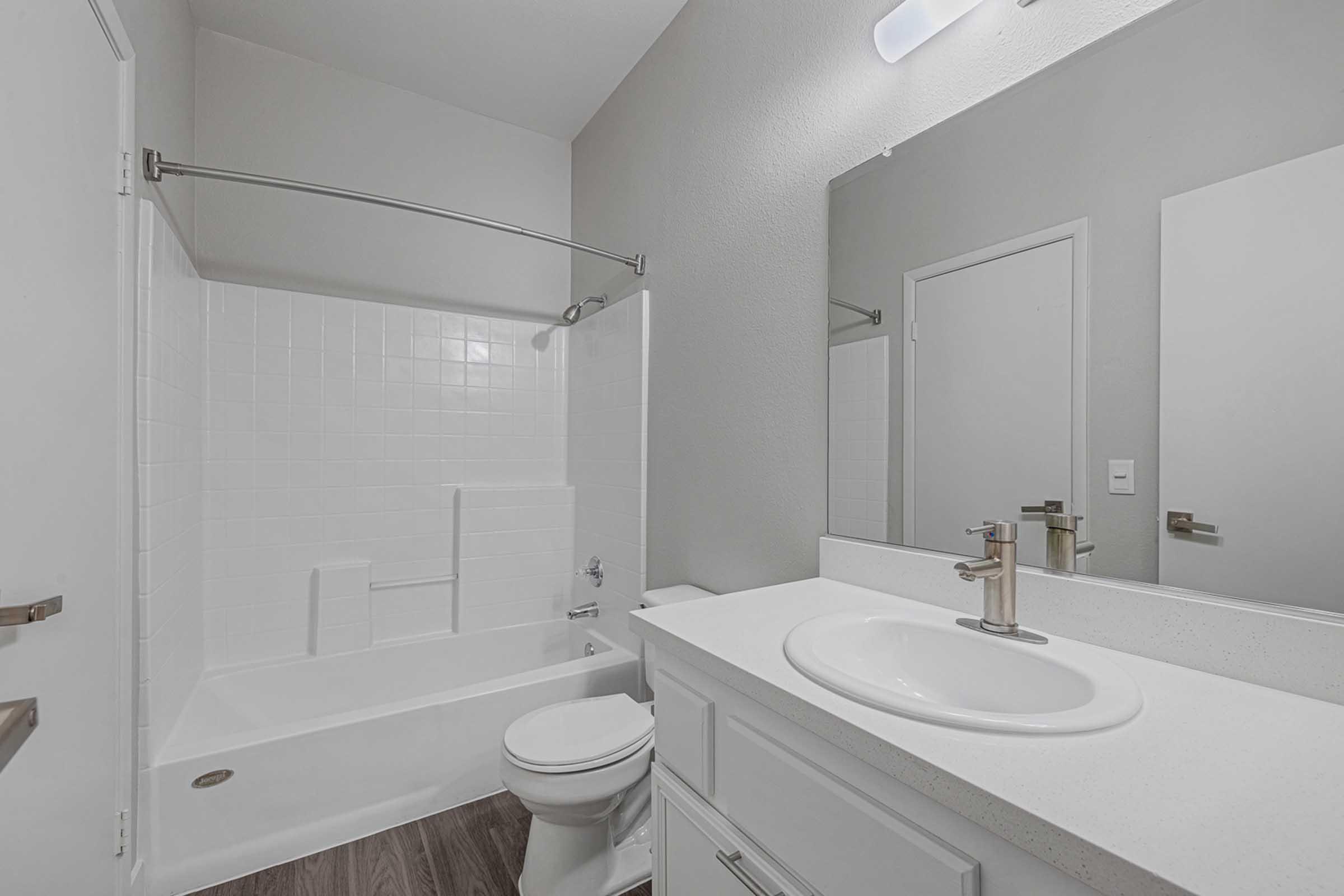 Modern bathroom featuring a white bathtub with a showerhead, a white countertop with a circular sink, and a sleek faucet. The walls are painted in a light gray tone, and there is a large mirror above the sink. A towel bar is mounted on the wall, and the floor has a wood-like finish.
