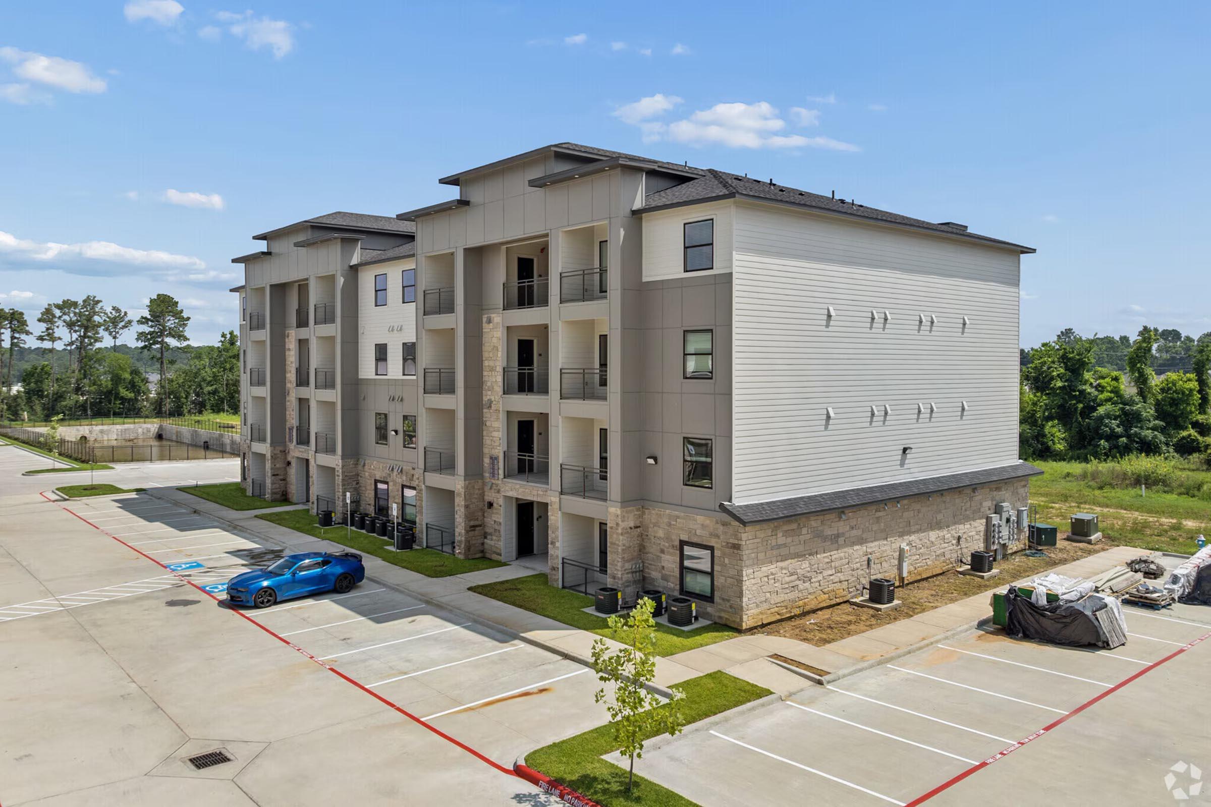 A modern multi-story apartment building with a light-colored exterior, featuring balconies and stone accents. The building is surrounded by green landscaping and parking spaces, including a blue car parked nearby. In the background, there are trees and open fields under a clear blue sky.
