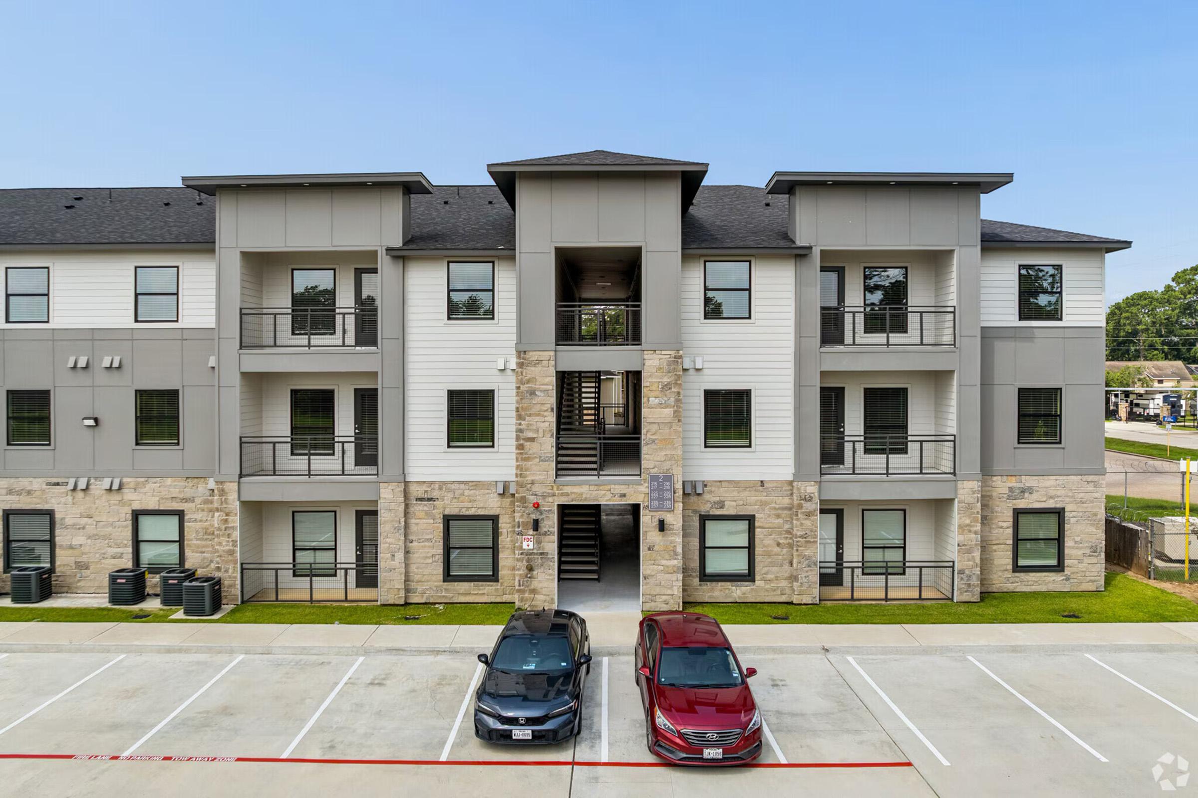 A modern three-story apartment building featuring a mix of white and gray siding with stone accents. The exterior includes balconies and multiple staircases. In the foreground, two parked cars are visible on a spacious paved area with several empty parking spaces. The sky is clear and blue.