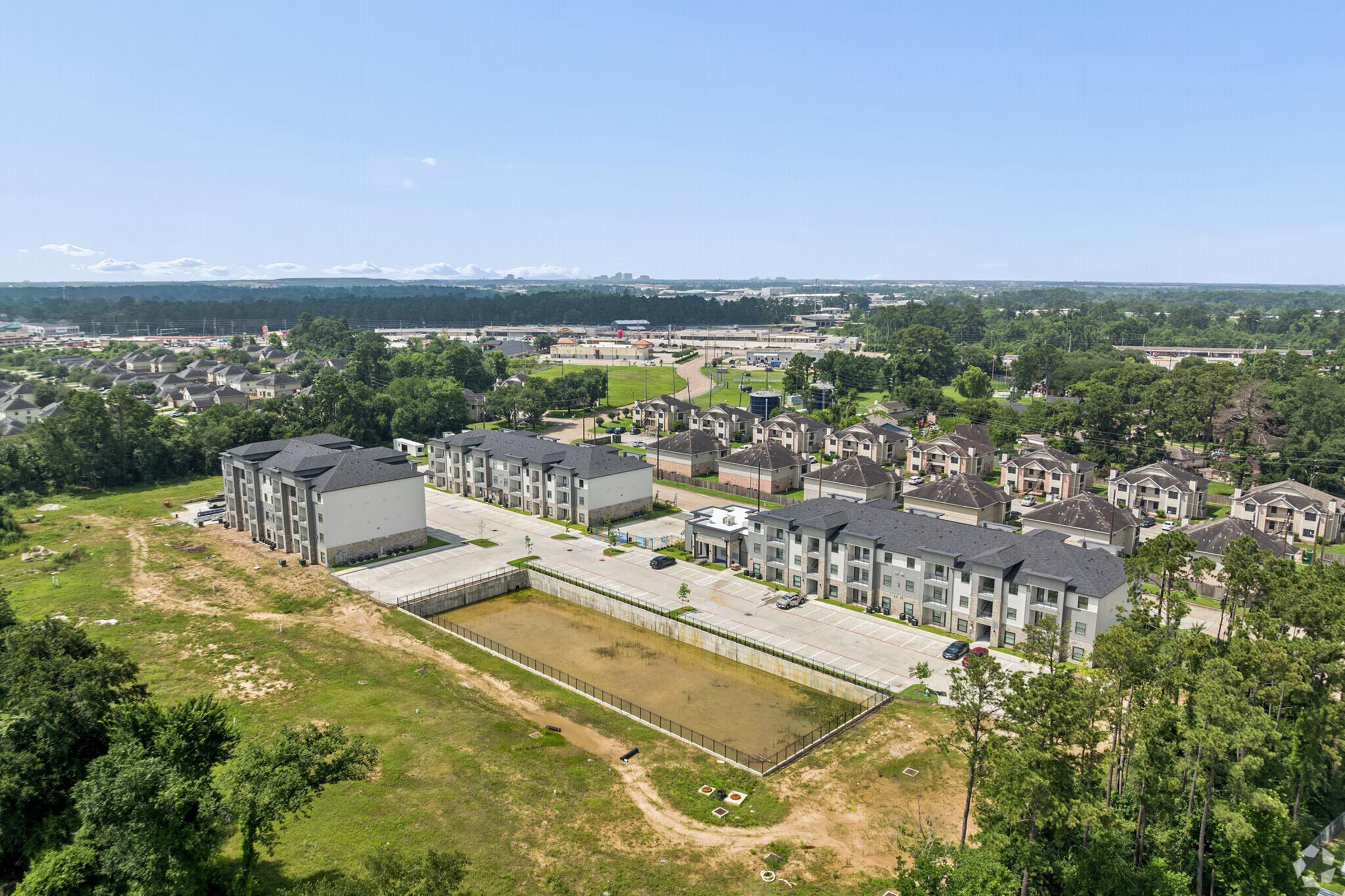 Aerial view of a residential area featuring several multi-story apartment buildings, a parking lot, and a small pond. Surrounding the development are green trees and open fields, with a distant view of highways and commercial structures beyond the apartments. Clear blue sky above.