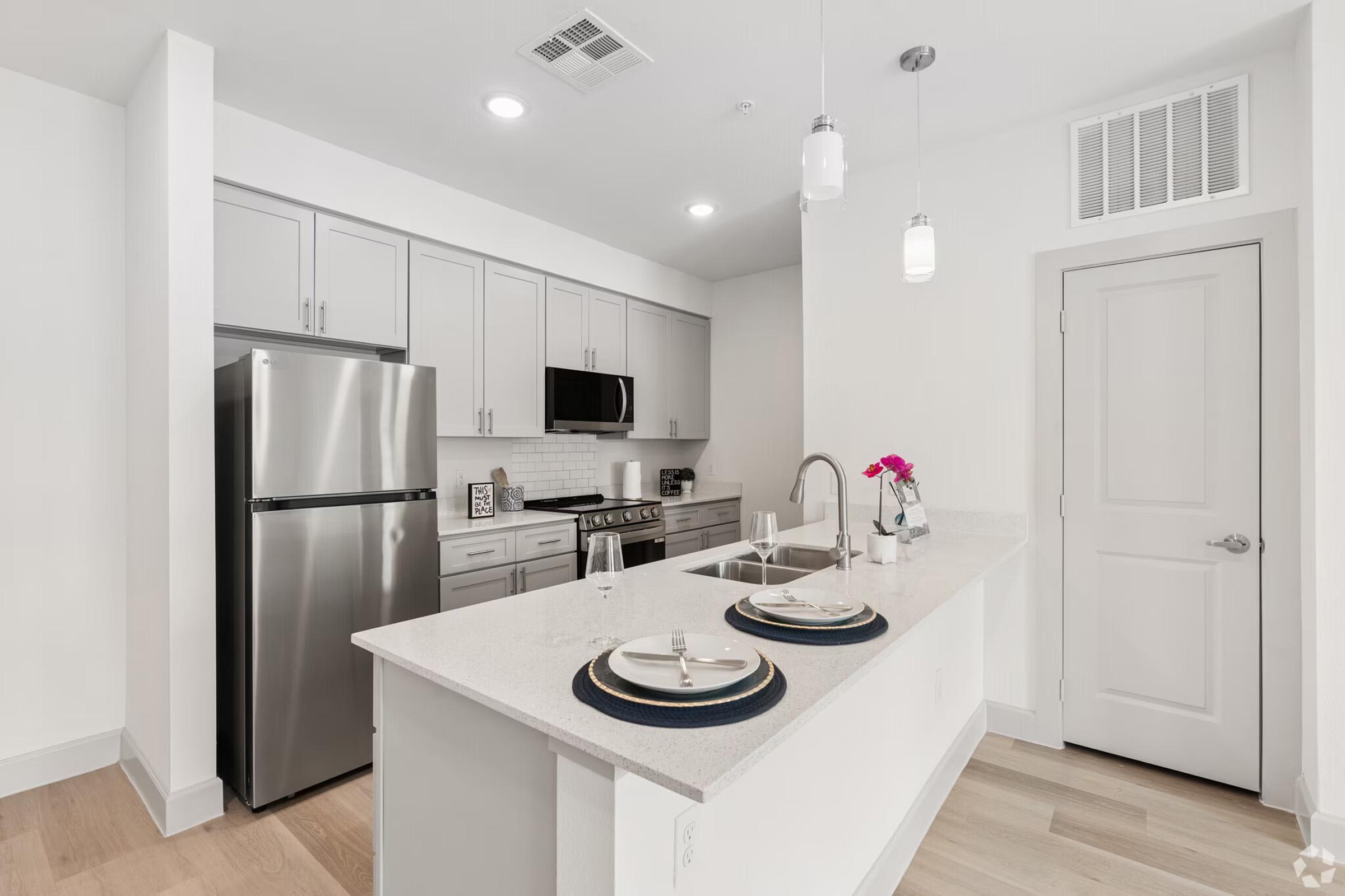 Modern kitchen featuring stainless steel appliances, including a refrigerator and microwave. A sleek countertop with two plates and glasses is set on a kitchen island. Light cabinetry complements the bright, open space, illuminated by pendant lighting. A door leads to another room, adding to the contemporary design.