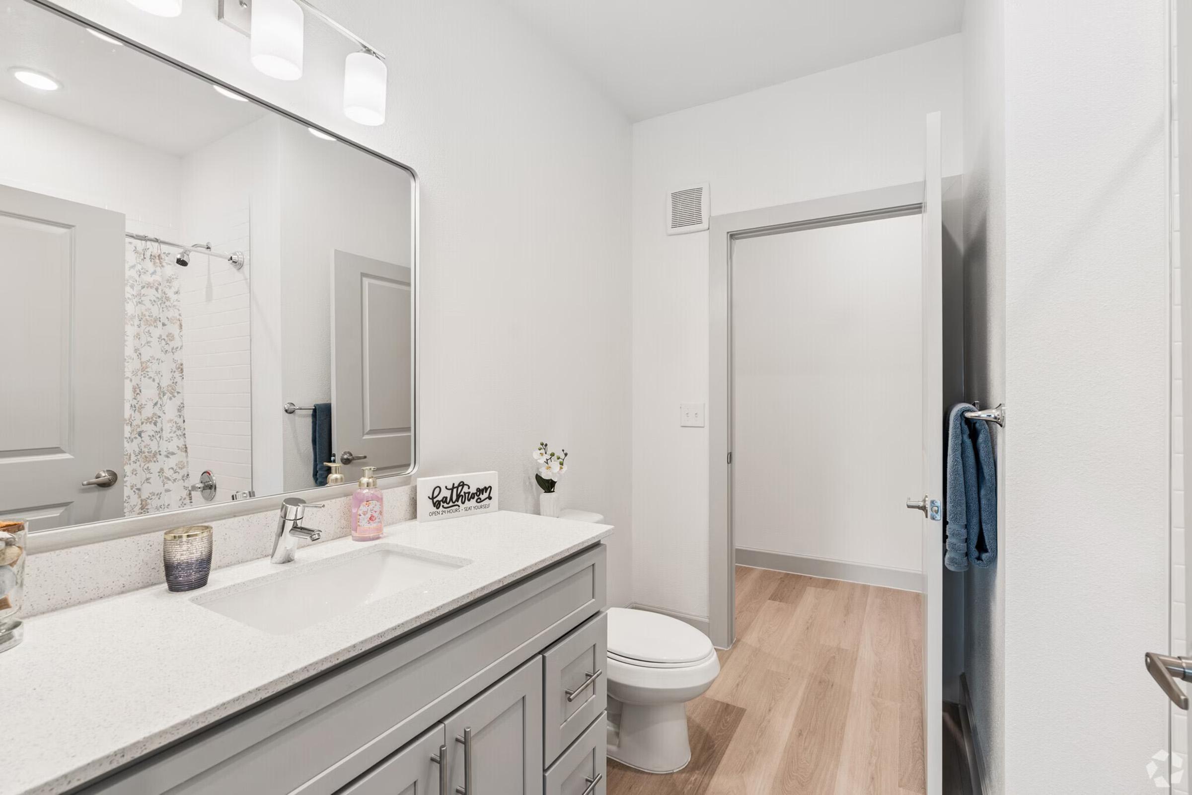 A modern bathroom featuring a double sink vanity with a large mirror above. The countertop is white with decorative items. There’s a light-colored tile shower with a floral curtain in the background, and a white toilet. A door leads to a hallway, and a blue towel hangs on the wall. The flooring is light wood.
