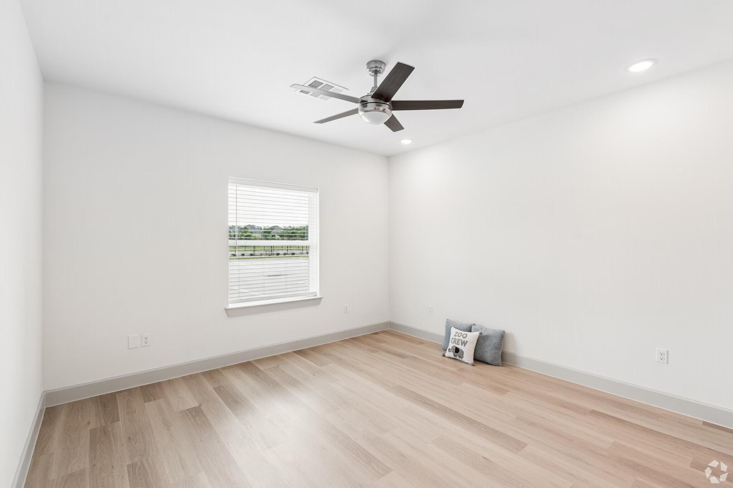 A bright, empty room with light-colored hardwood flooring, white walls, and a ceiling fan. A window allows natural light to enter, revealing a view outside. In one corner, there is a small decorative pillow with the words "You Are Loved." The space is minimalistic and modern, perfect for personal customization.