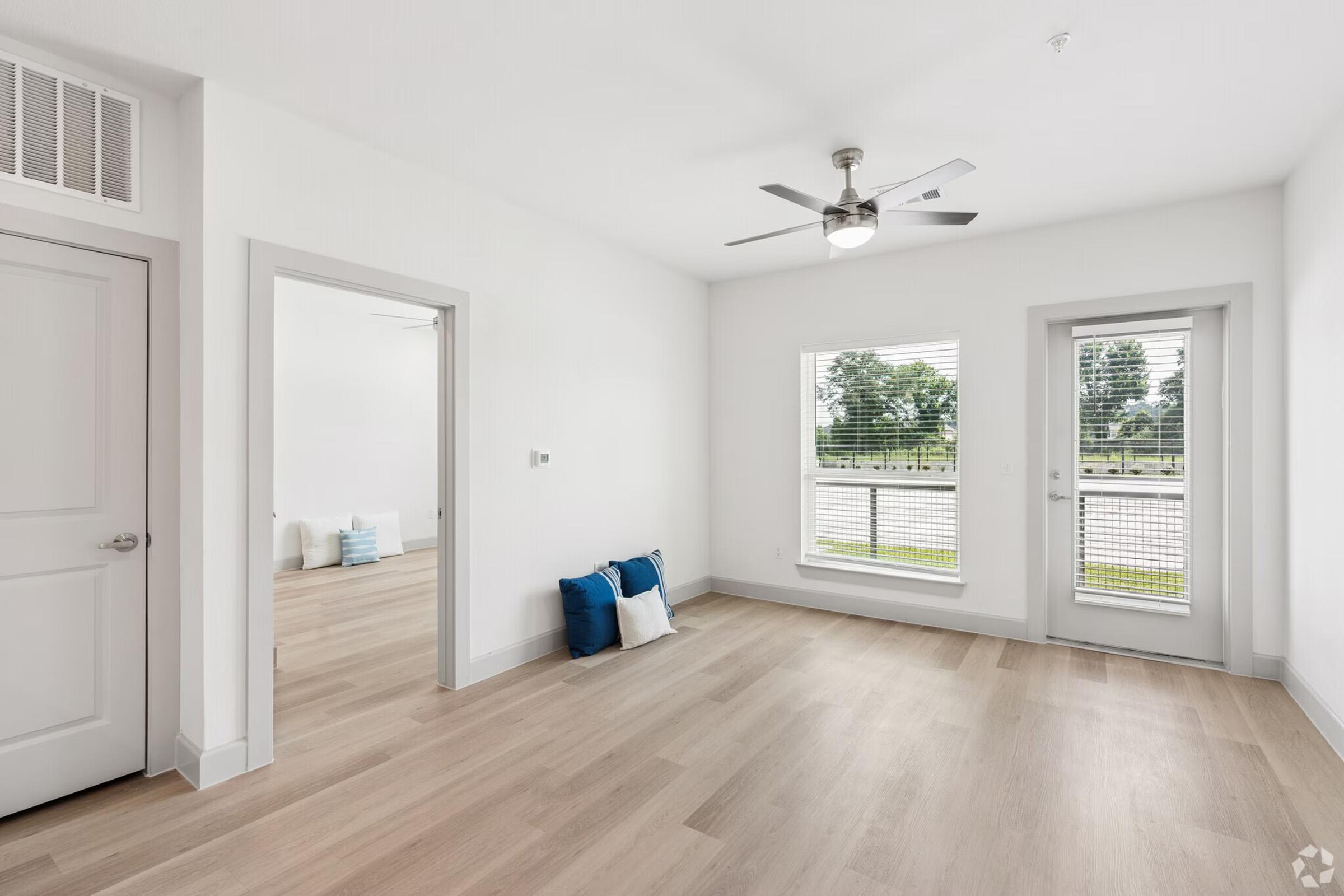 A bright, modern living space with light-colored walls and wooden flooring. The room features a ceiling fan and two large windows with blinds, providing natural light. In the corner, there are decorative pillows in blue and white. A doorway leads to another area of the space.