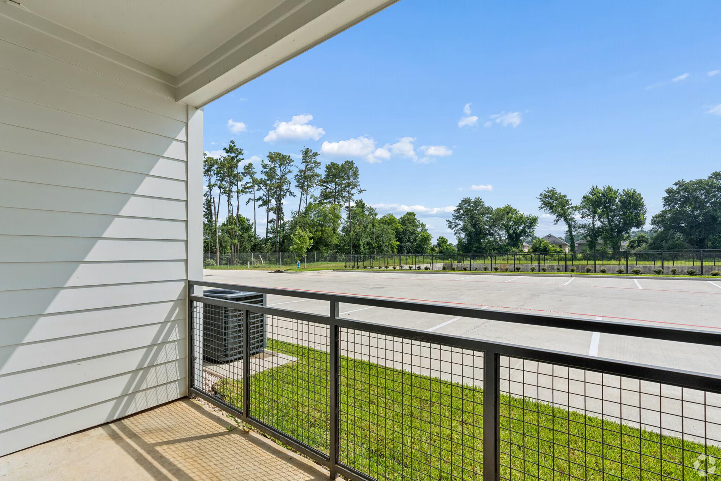 View from a balcony, showcasing a clear sky and green trees in the background. An empty parking lot is visible below, with a fence surrounding the area. The balcony features a metal railing and is partially shaded by the building.