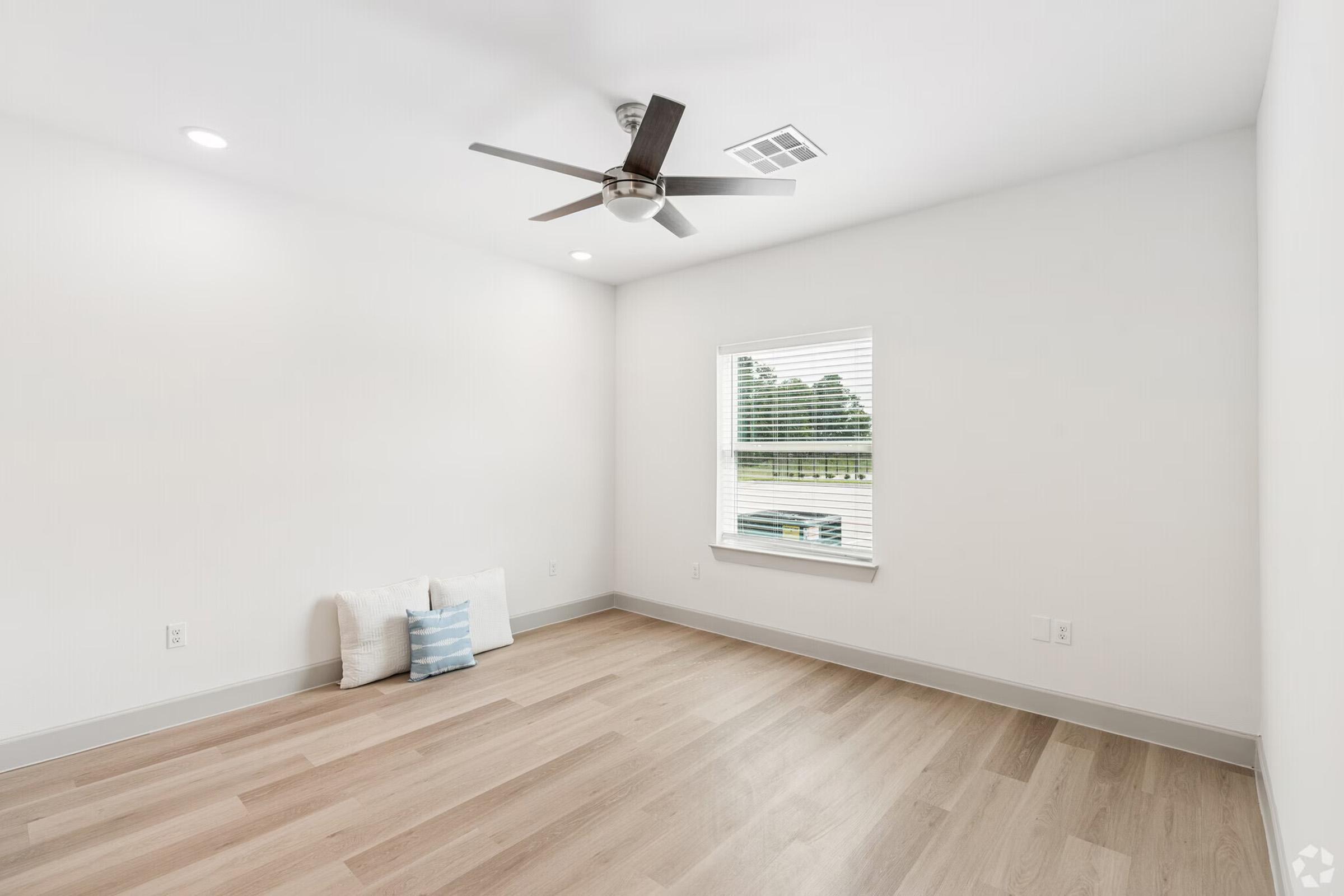 A bright, empty room with light-colored walls and a hardwood floor. There is a ceiling fan and a window with white blinds, allowing natural light to enter. Two decorative cushions are placed on the floor near the window, creating a minimalist and modern aesthetic.