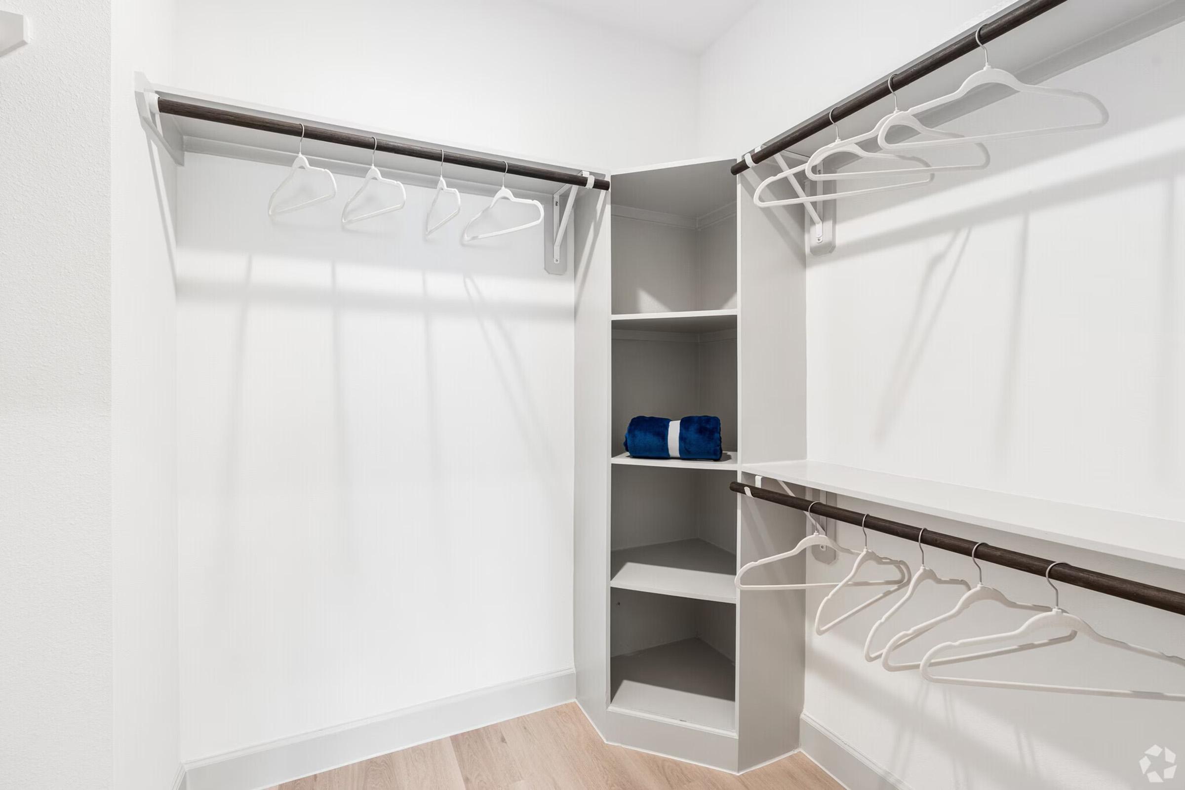 A spacious, organized closet featuring several empty hangers on two rods, a set of neatly folded blue towels on a shelf, and an open corner shelf. The design includes bright white walls and light wood flooring, creating a clean and airy atmosphere.