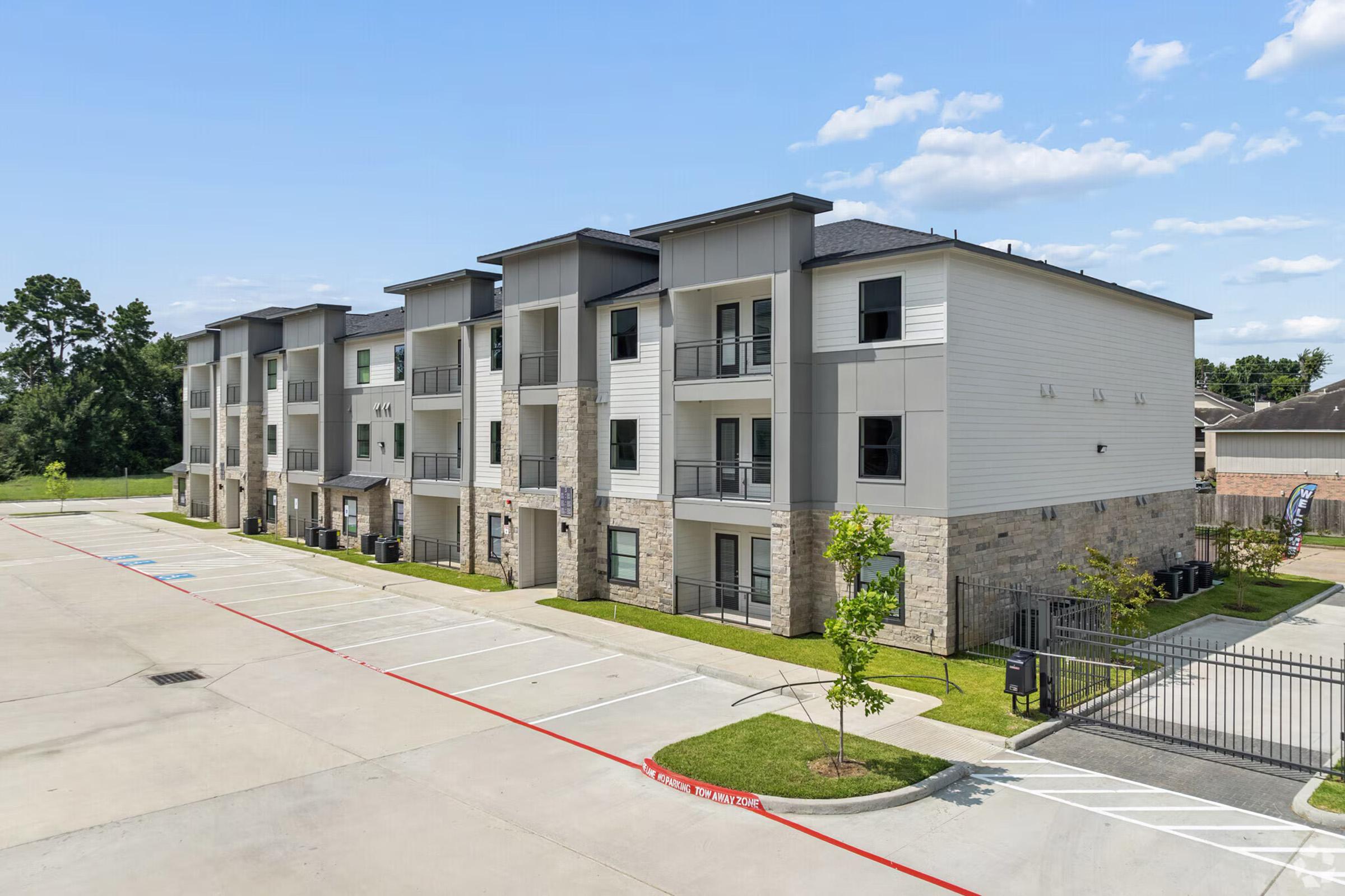 A modern multi-story apartment building featuring a combination of light-colored siding and stone accents. The building is set in a spacious parking lot with designated parking spaces and small trees. Clear blue skies provide a bright backdrop for the scene.