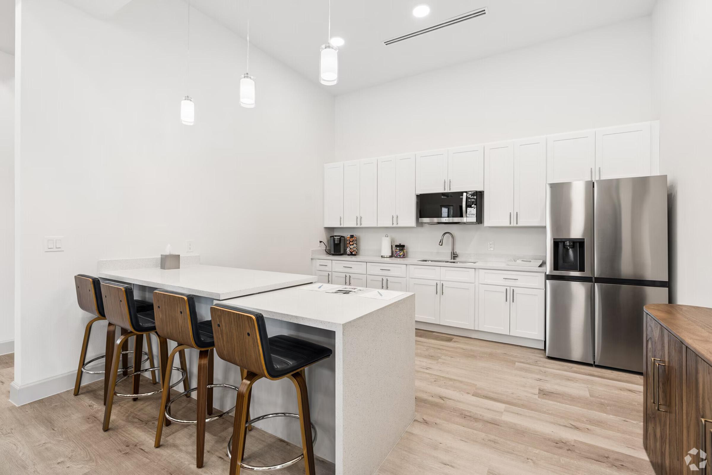 Modern kitchen featuring white cabinetry, stainless steel appliances including a refrigerator and microwave, and a spacious island with seating for four. The flooring is light wood, and the space is well-lit with hanging light fixtures. A minimalist design creates a clean, contemporary look.