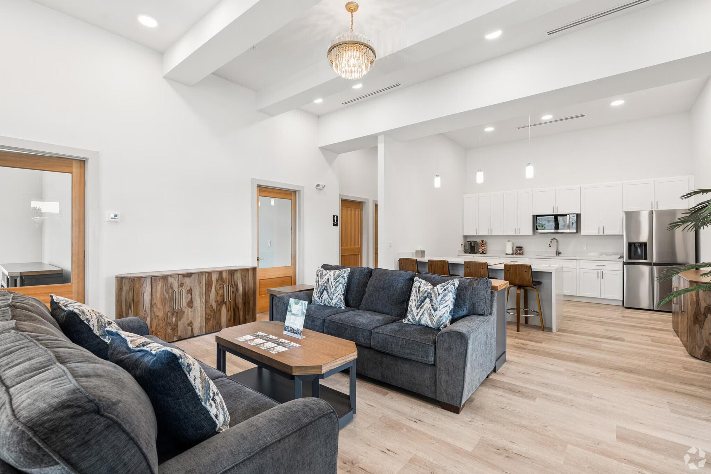 A spacious, modern living area featuring a dark grey sectional sofa with decorative pillows, a wooden coffee table, and a large kitchen counter with bar stools. The room has bright lighting from overhead fixtures and large windows, with a contemporary aesthetic and light wooden flooring.