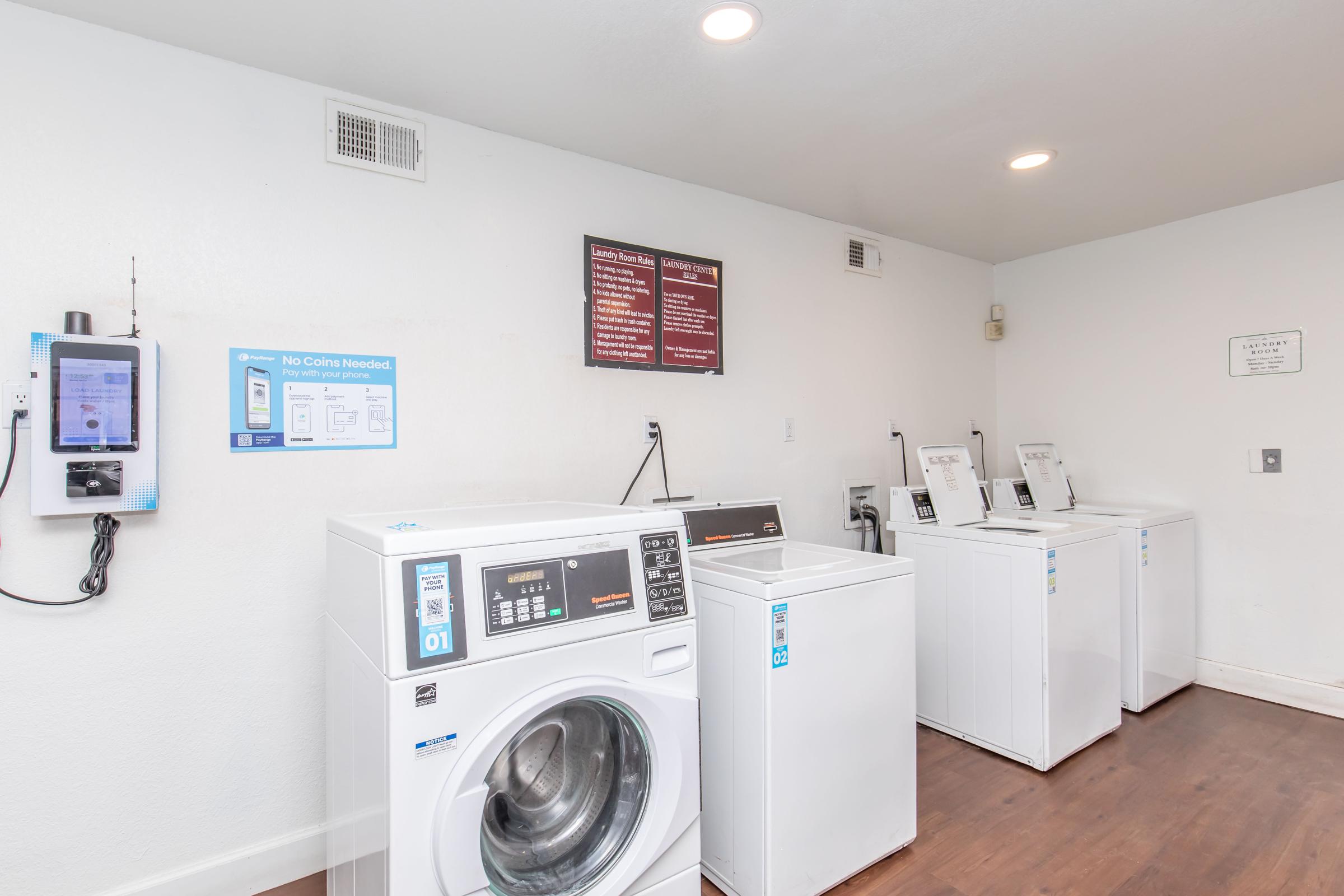 Laundry room featuring several white washing machines and dryers. A payment machine is visible on the left. There are instructional signs on the wall regarding laundry services. The space has a clean, well-lit environment with wooden flooring.